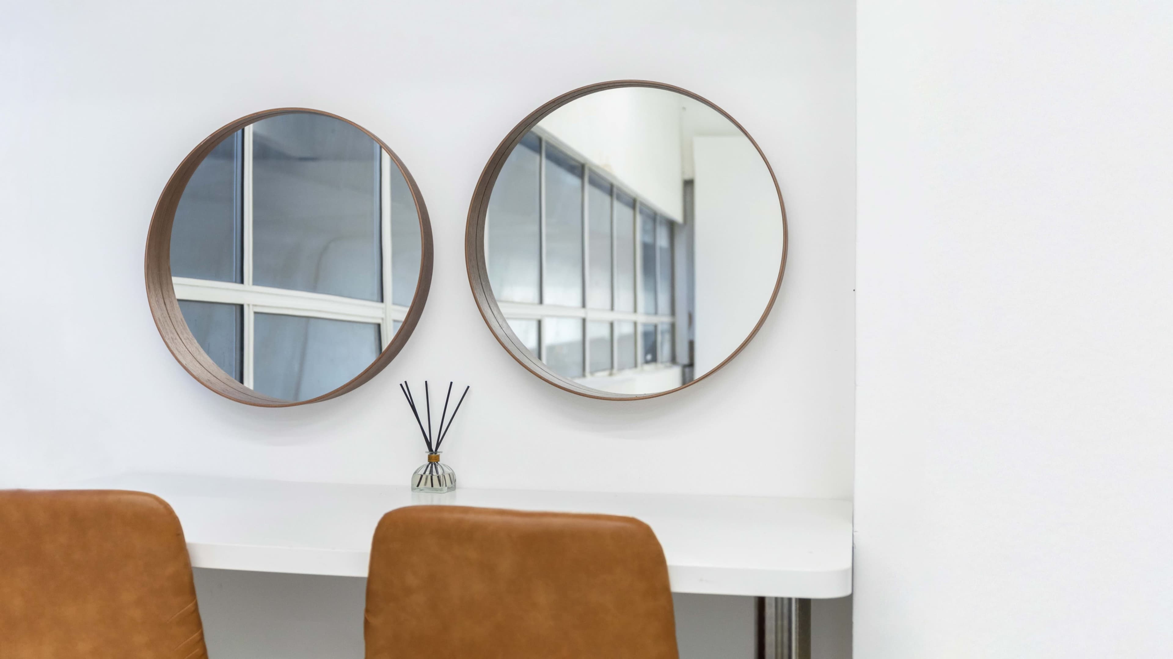 Two round mirrors are mounted on a white wall above a light-colored table with two brown chairs and a small decorative vase with reed sticks.