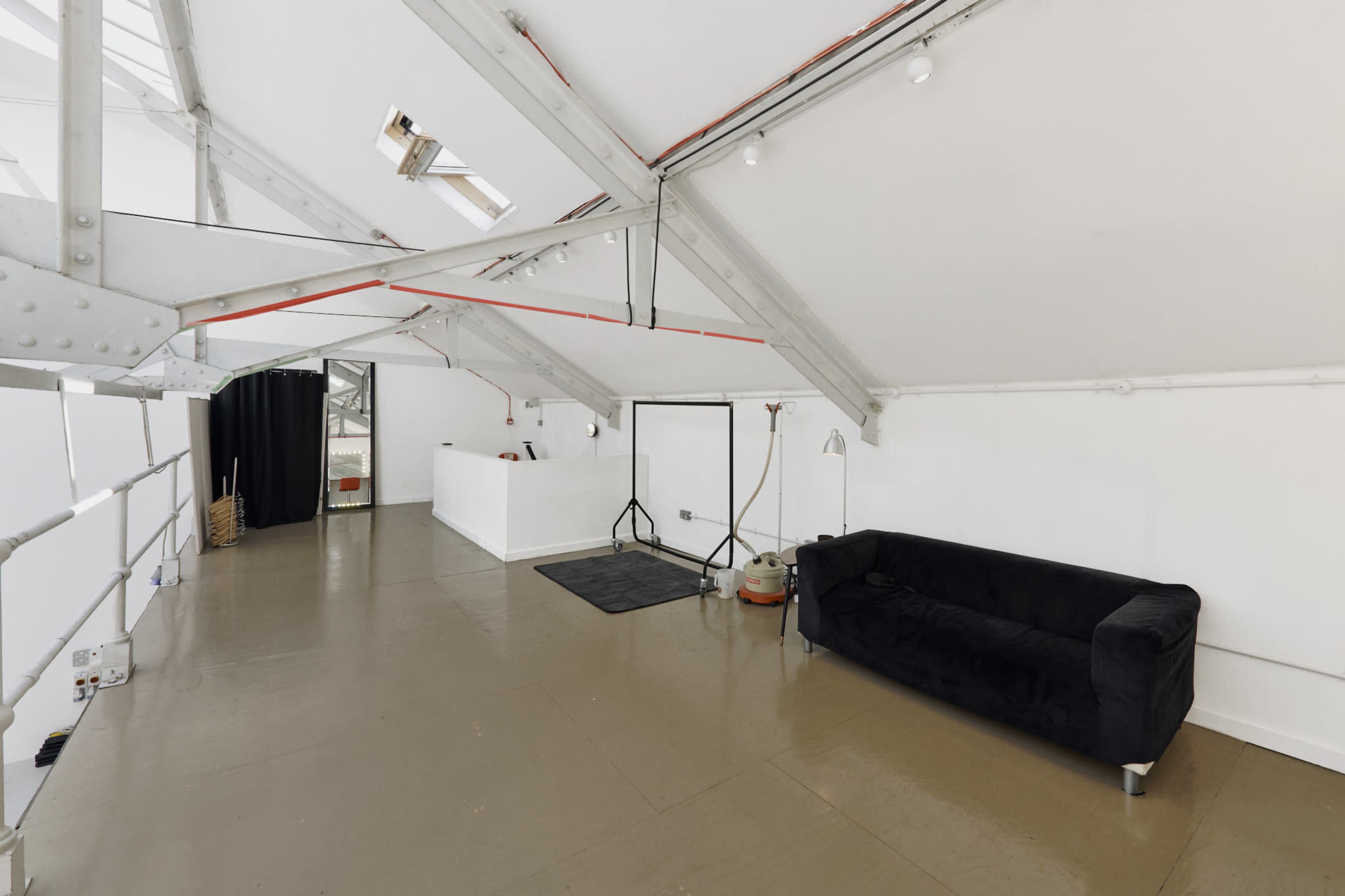 The image shows a spacious, bright room with a black couch, white walls, and exposed beams, featuring a few minimal furnishings and a large window.