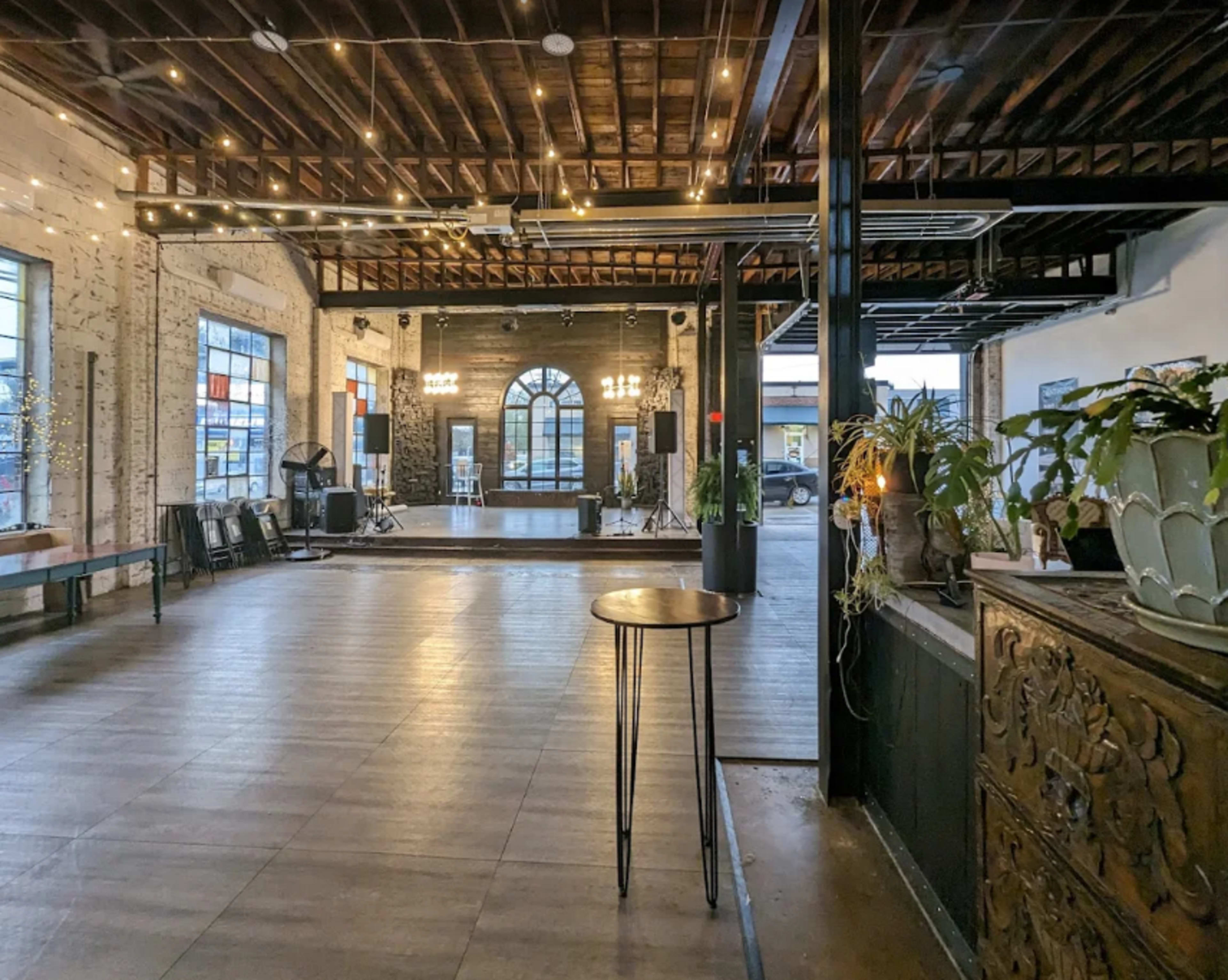 The image shows a spacious interior with wooden beams, large windows, and a polished floor, featuring a mix of decorative plants and furniture.