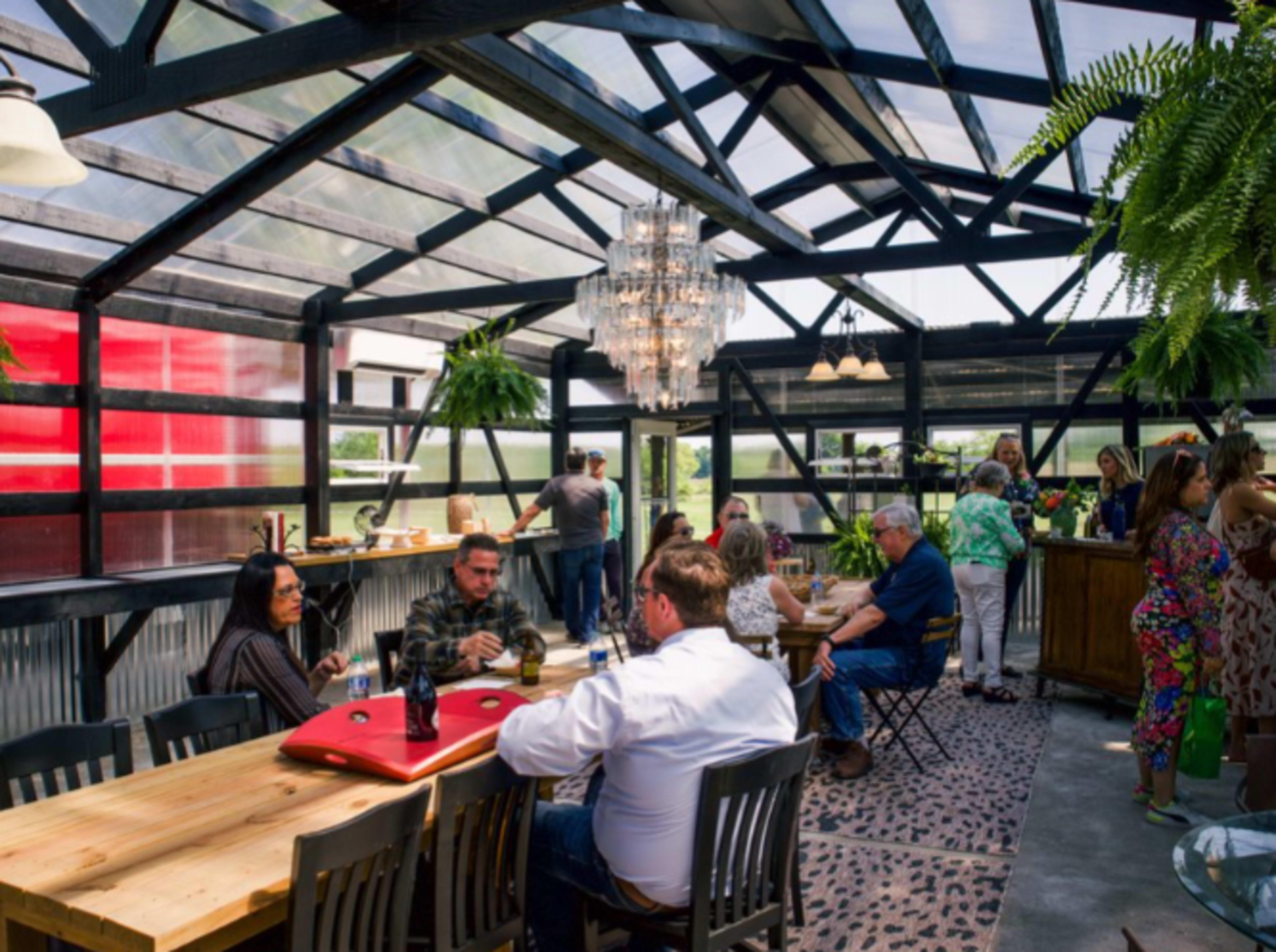 A bright, spacious indoor greenhouse with a dining area, featuring wooden tables, a chandelier, and patrons engaged in conversation.