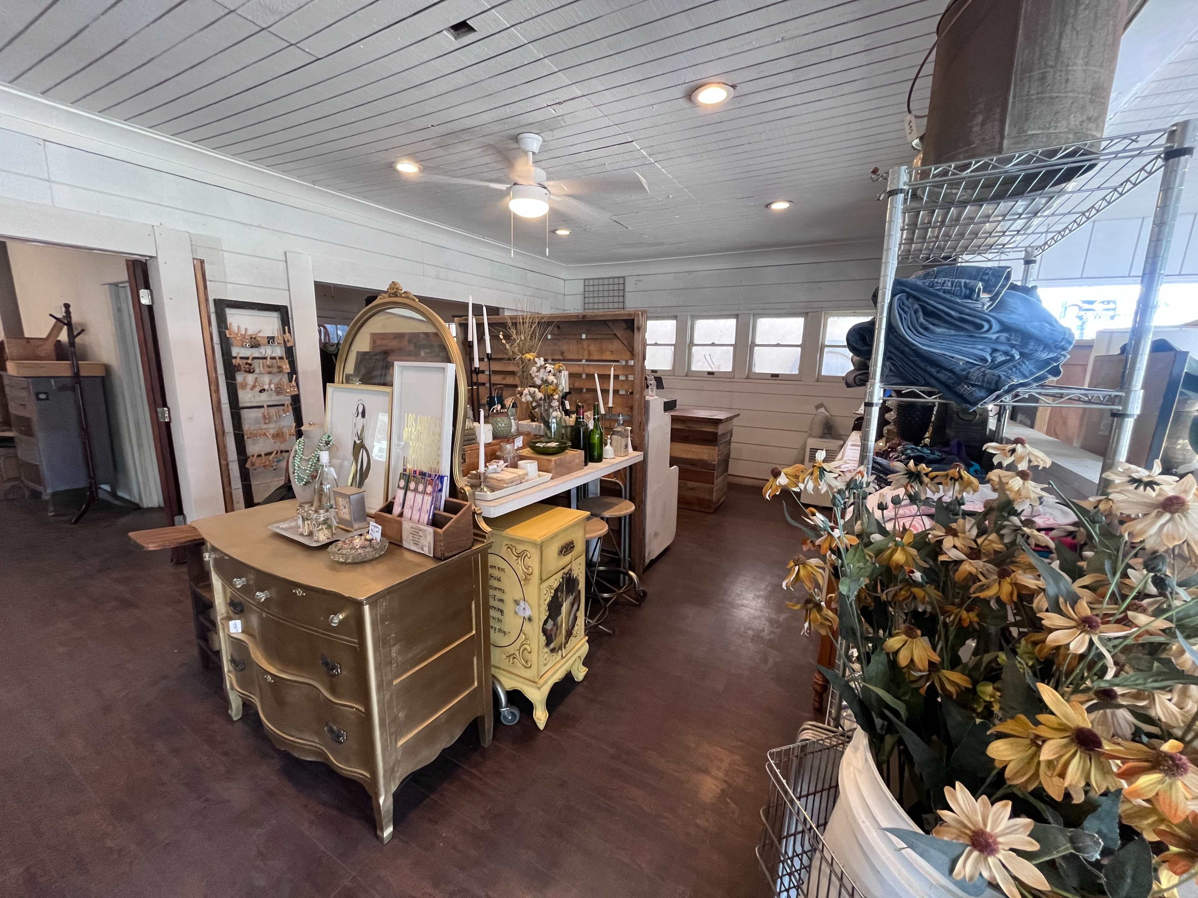 The image shows an interior space of a vintage shop featuring a gold-colored dresser, decorative items on a table, shelves, and floral arrangements.
