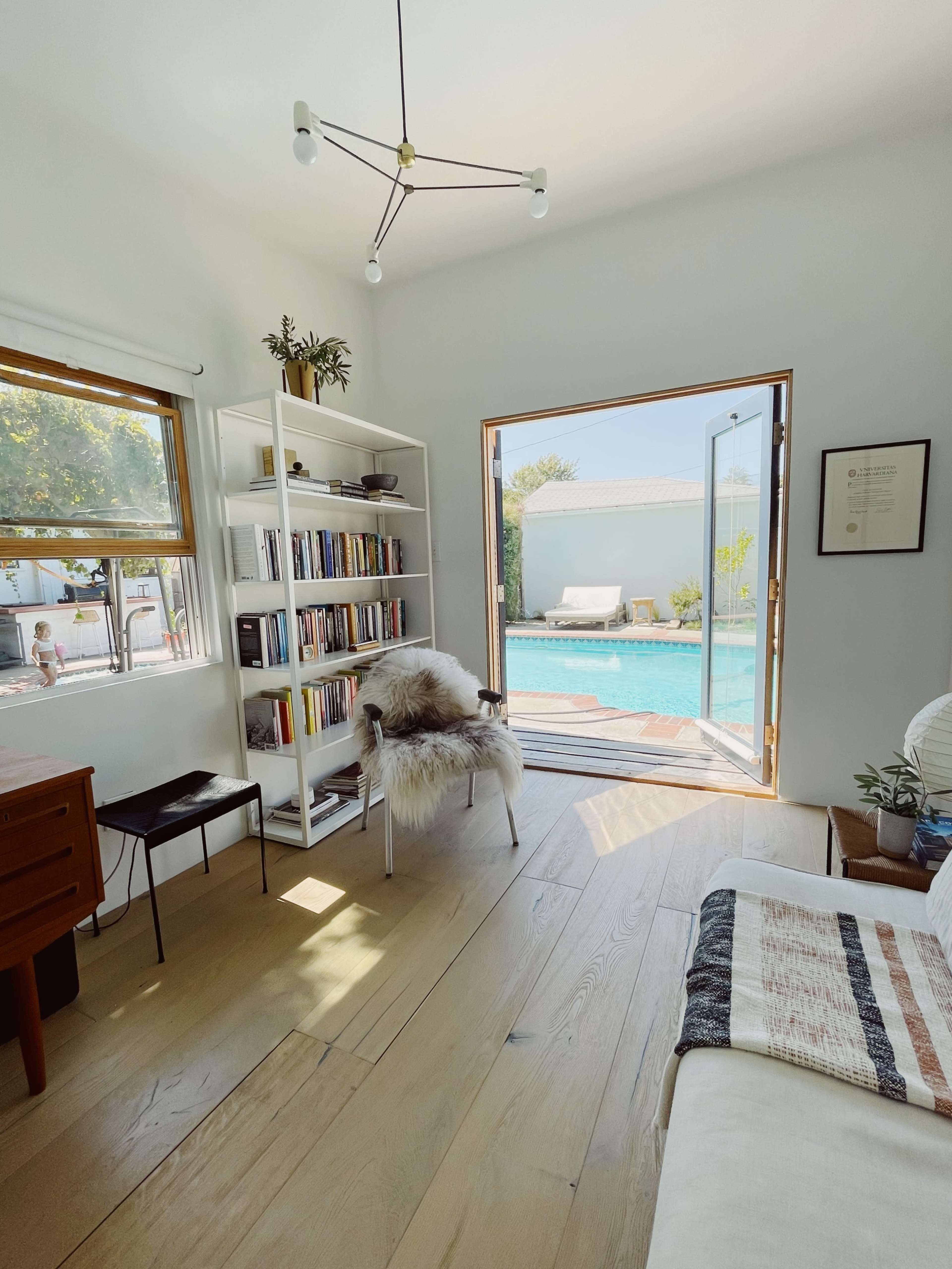 A bright room features a bookshelf and large open doors that lead to a pool area outside.