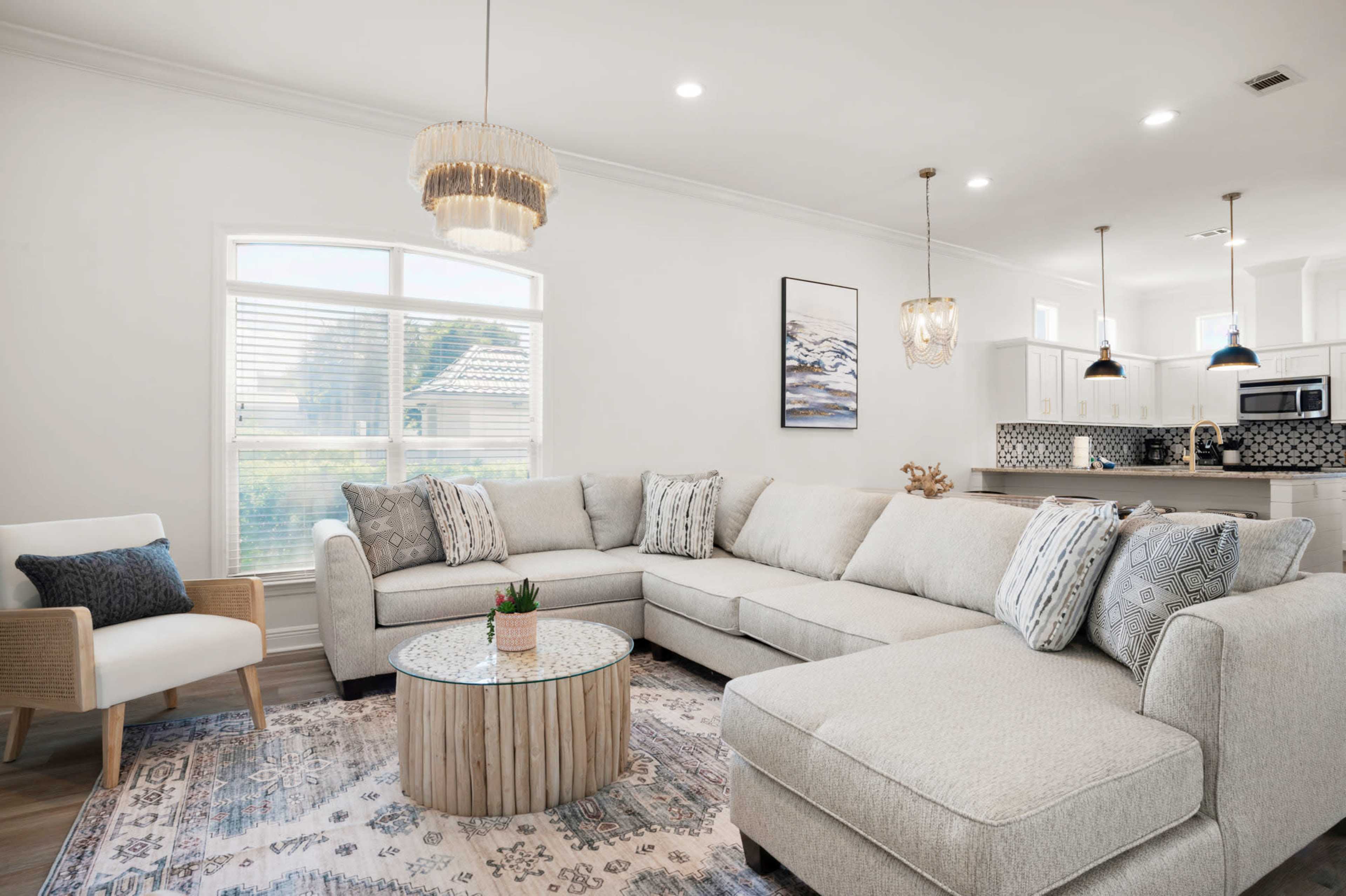 A light-colored living room featuring a large sectional sofa, a round wooden coffee table, and a modern decorative rug, with pendant lights hanging above the space.