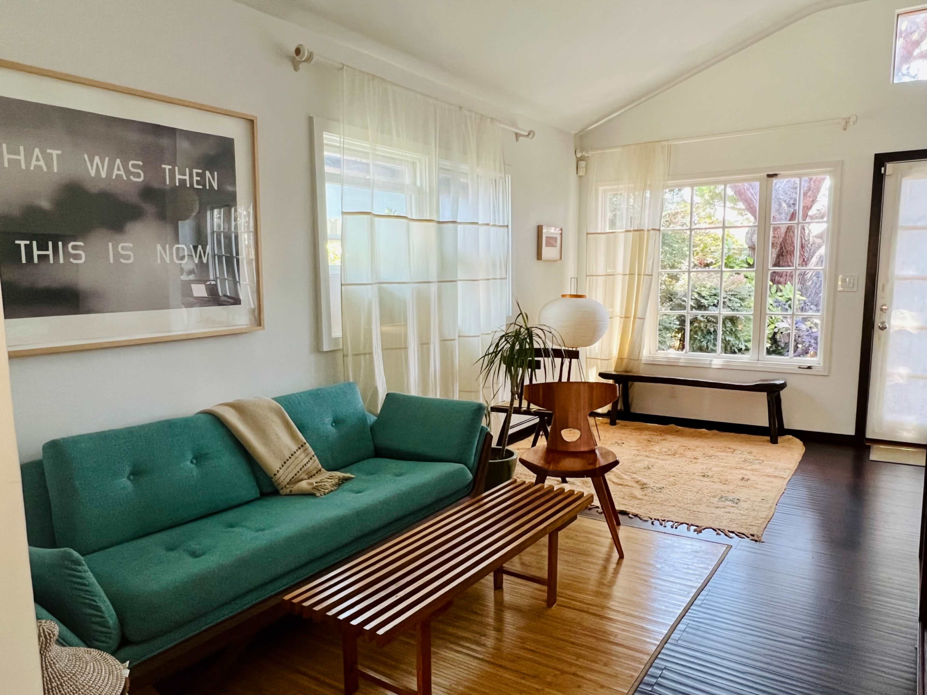 A bright living room featuring a teal sofa, a wooden coffee table, a floor lamp, and large windows with sheer curtains.