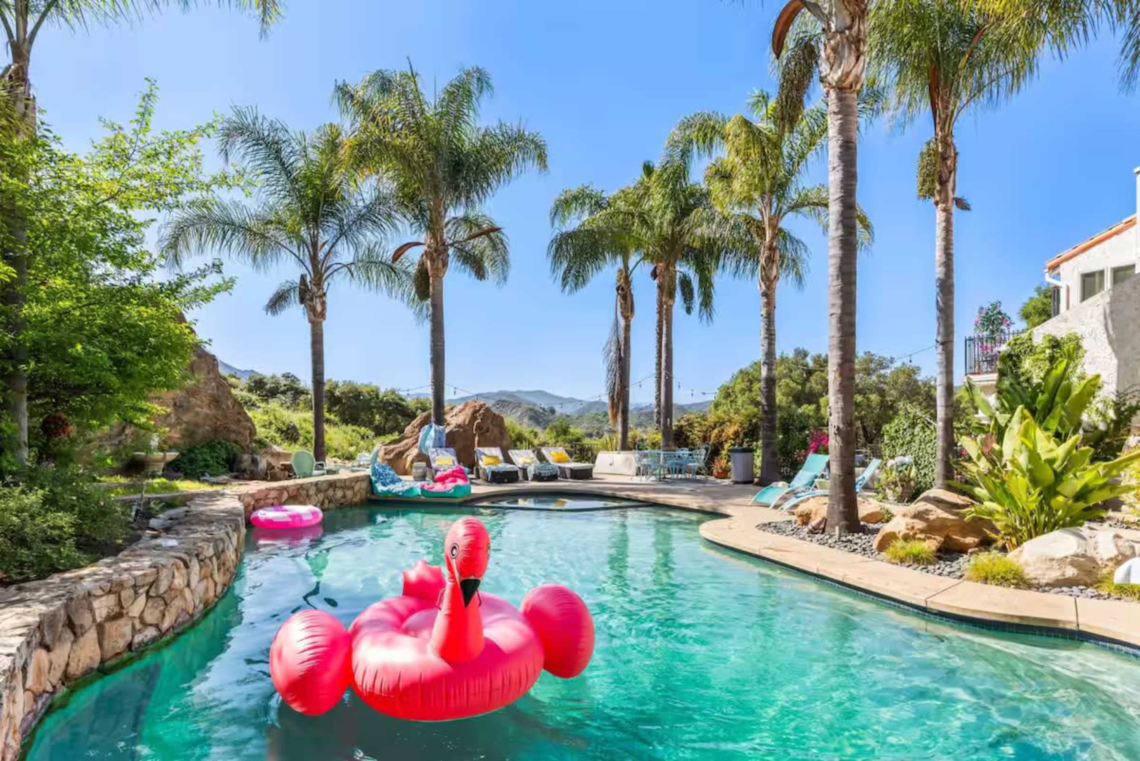 The image shows a swimming pool surrounded by palm trees and a rocky landscape, featuring a large inflatable pink flamingo floating on the water.