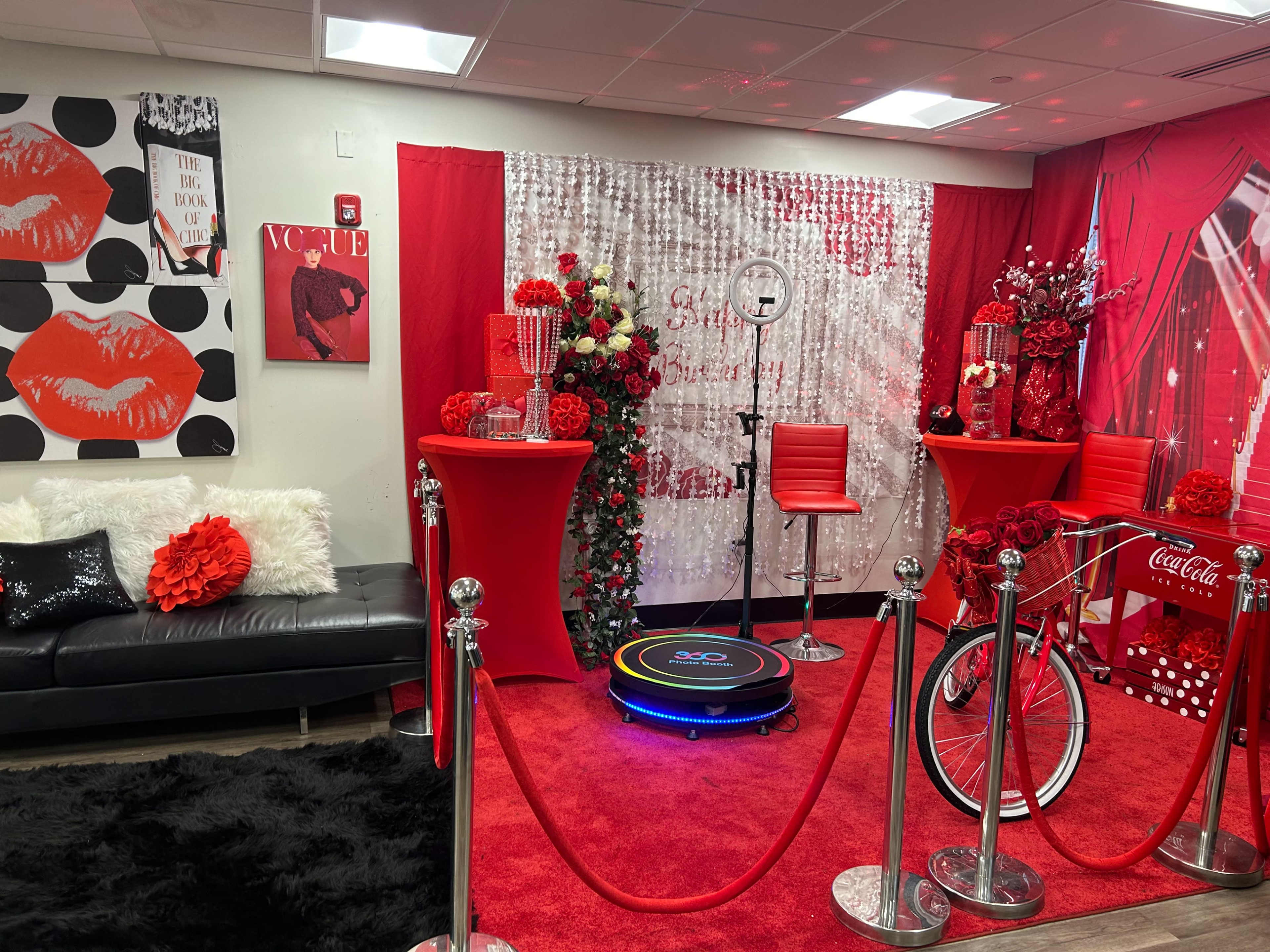 The scene features a vibrant event space adorned with red decorations, a black leather couch, and a vintage-style bicycle, all complemented by a shimmering backdrop and floral arrangements.