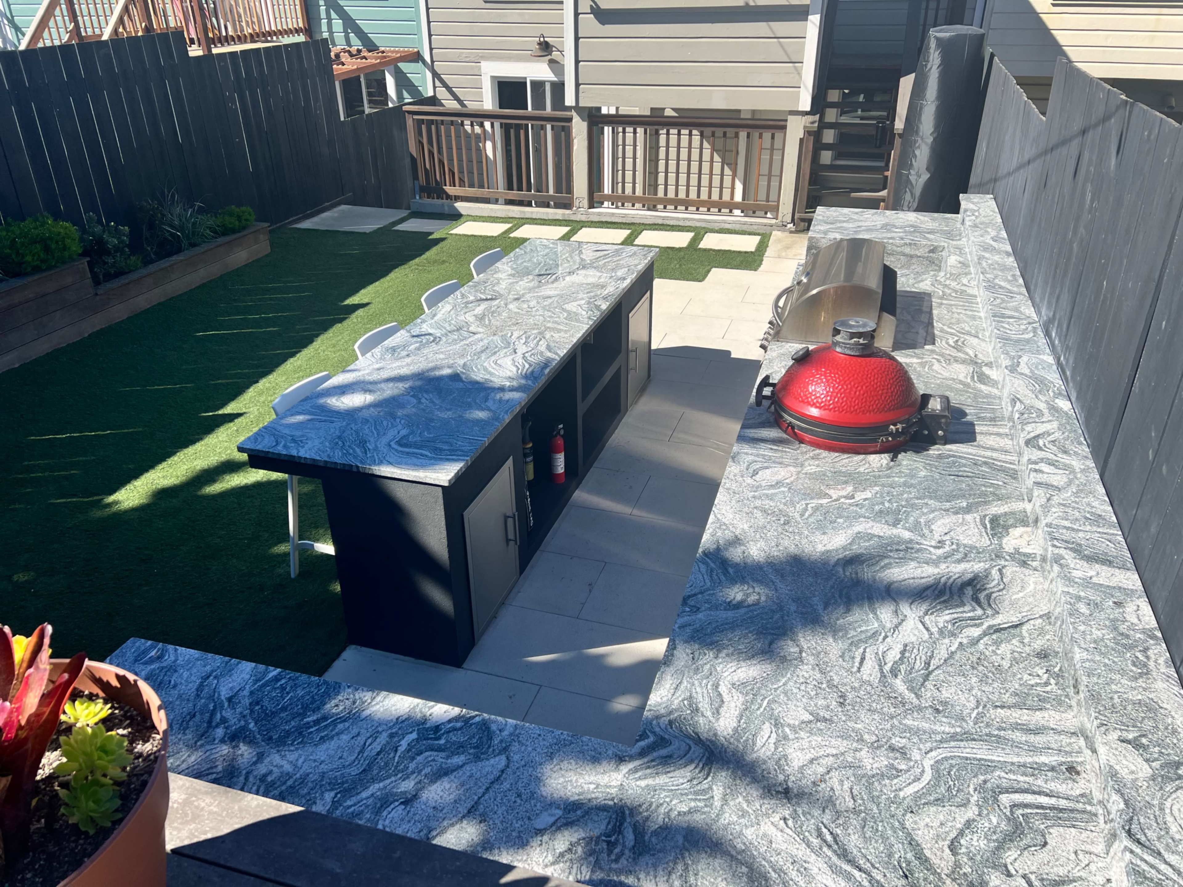 The image shows an outdoor kitchen area with a marble countertop, a red grill, and a green lawn surrounded by wooden fencing and nearby buildings.