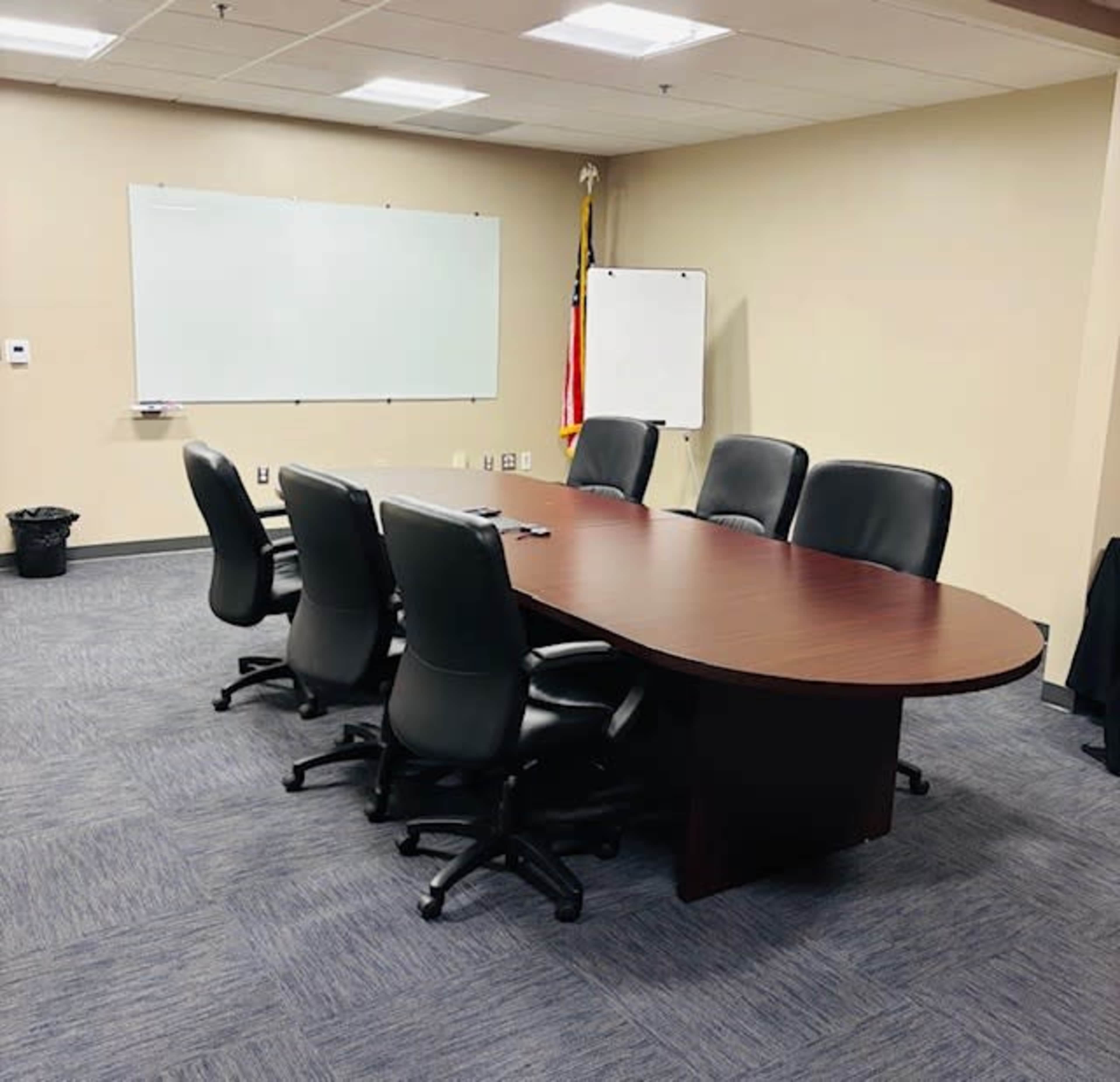 A conference room features a large oval table surrounded by six black chairs, with a whiteboard and a flip chart in the background.