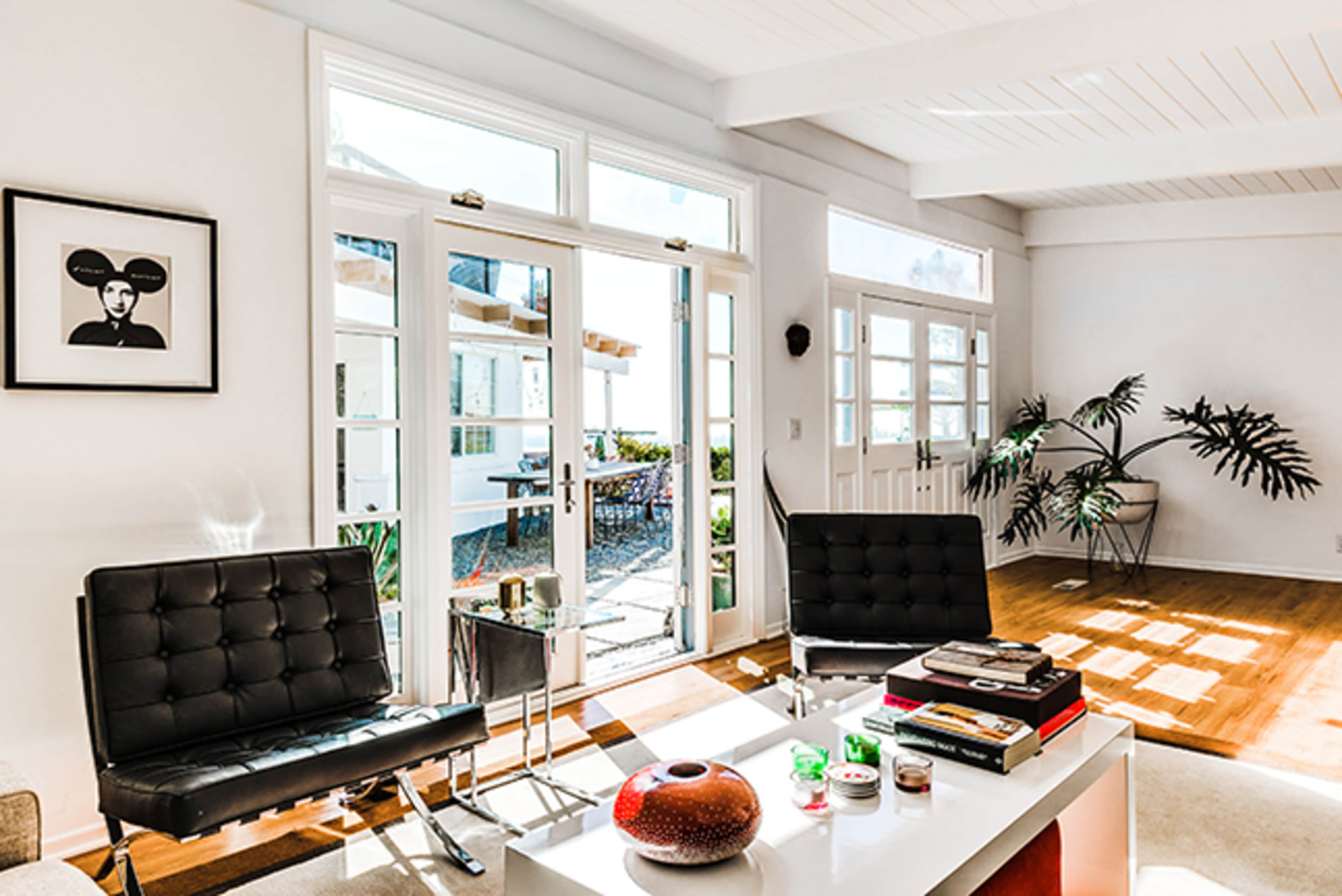 The image shows a bright, modern living room featuring two black lounge chairs, a coffee table with decorative items, and large glass doors that open to an outdoor patio.