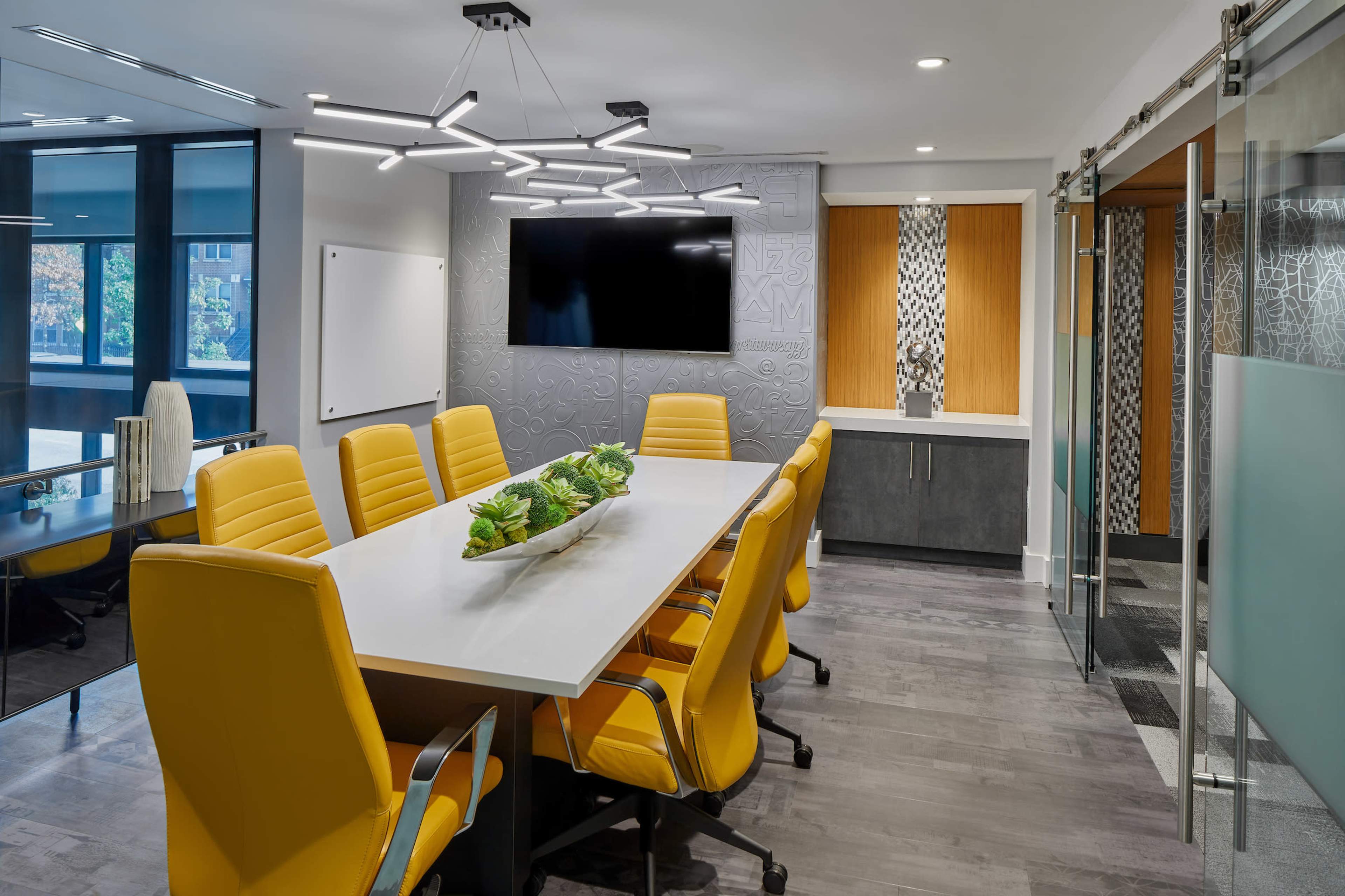 A modern conference room features a long table surrounded by yellow chairs, a wall-mounted TV, and large windows.