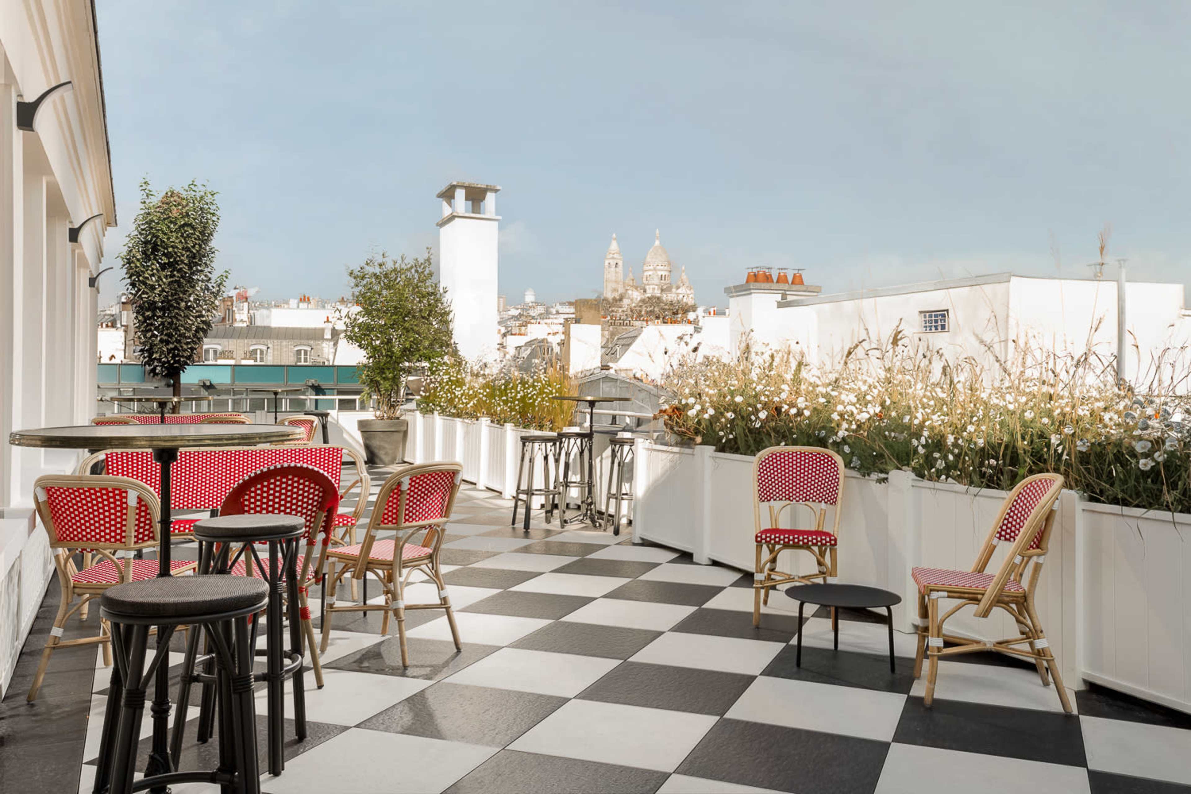 A rooftop terrace features black and white checkered flooring, surrounded by potted plants and chairs, with a distant view of a church dome.