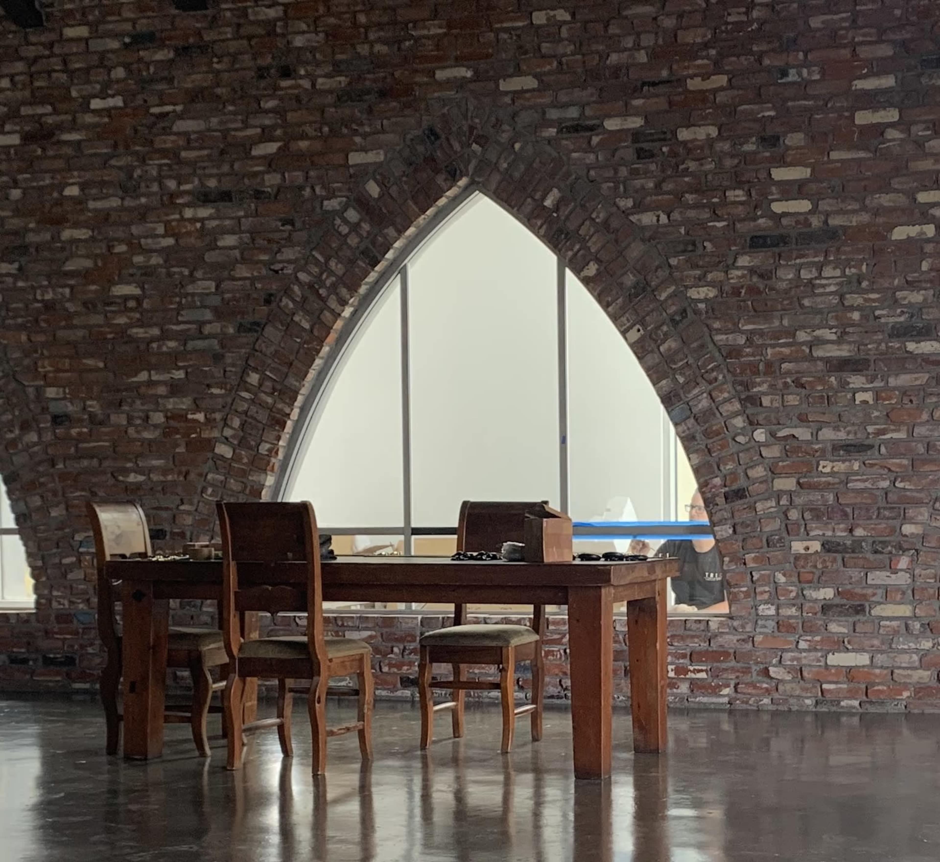 A wooden table and two chairs are positioned beside a large arched window in a room with a brick wall.