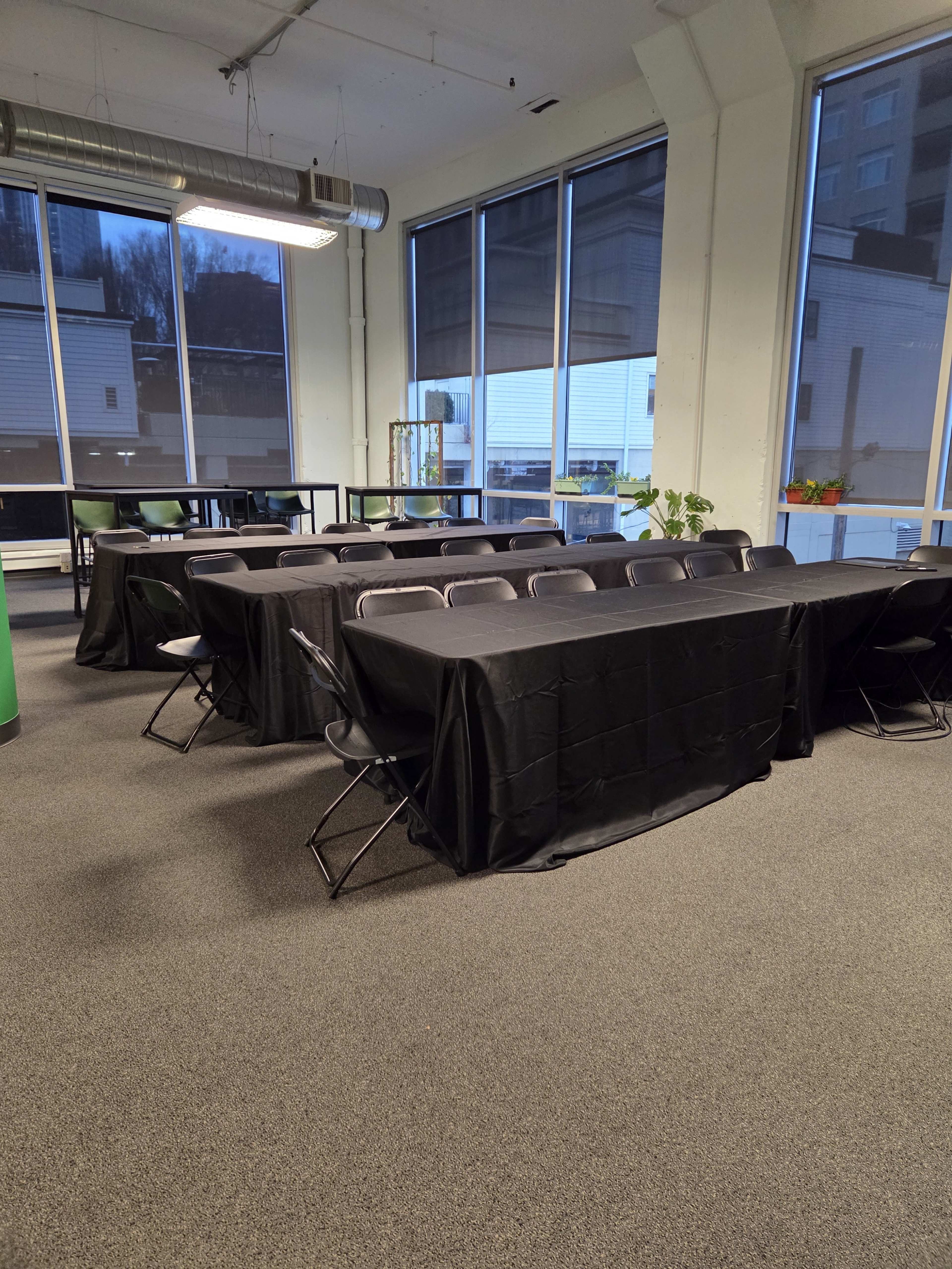 The image shows a room with several black-covered tables arranged in a U-shape, surrounded by folding chairs, and large windows letting in natural light.