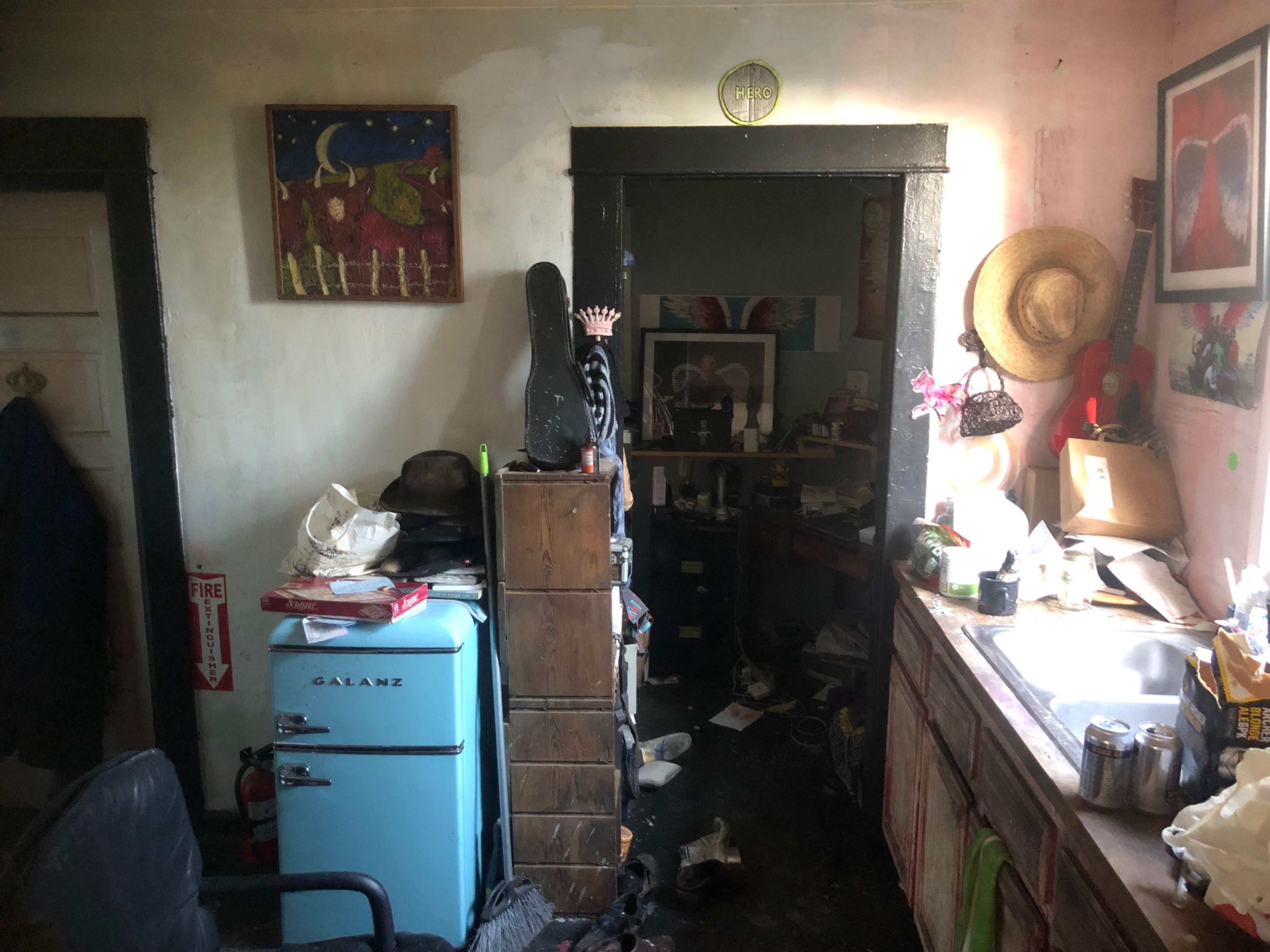 The image shows a cluttered room with a vintage blue refrigerator, a messy sink area, and dark furniture surrounded by disorganized personal items.