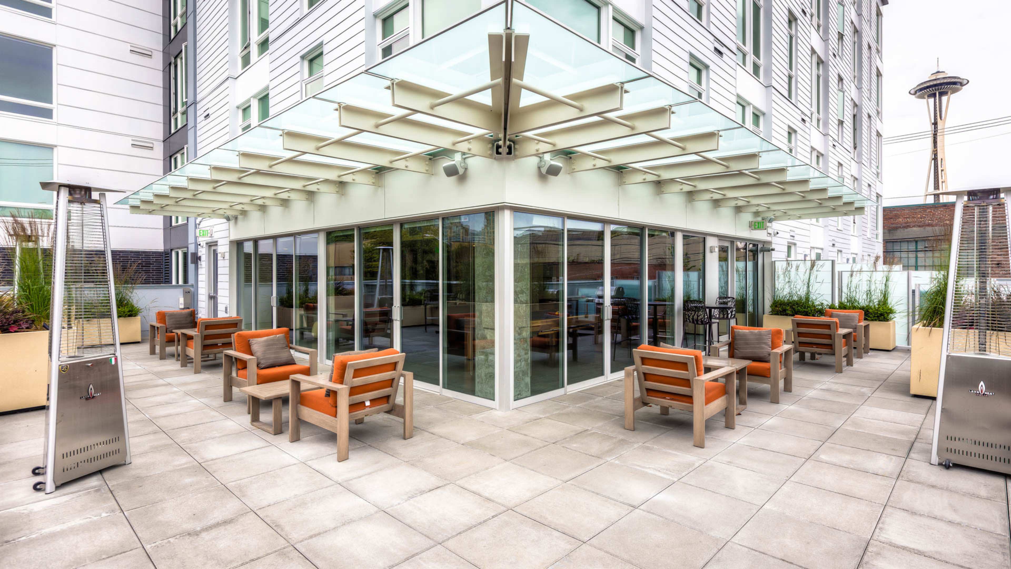 The image shows a modern outdoor seating area with metal and wooden furniture, surrounded by glass walls and planters.