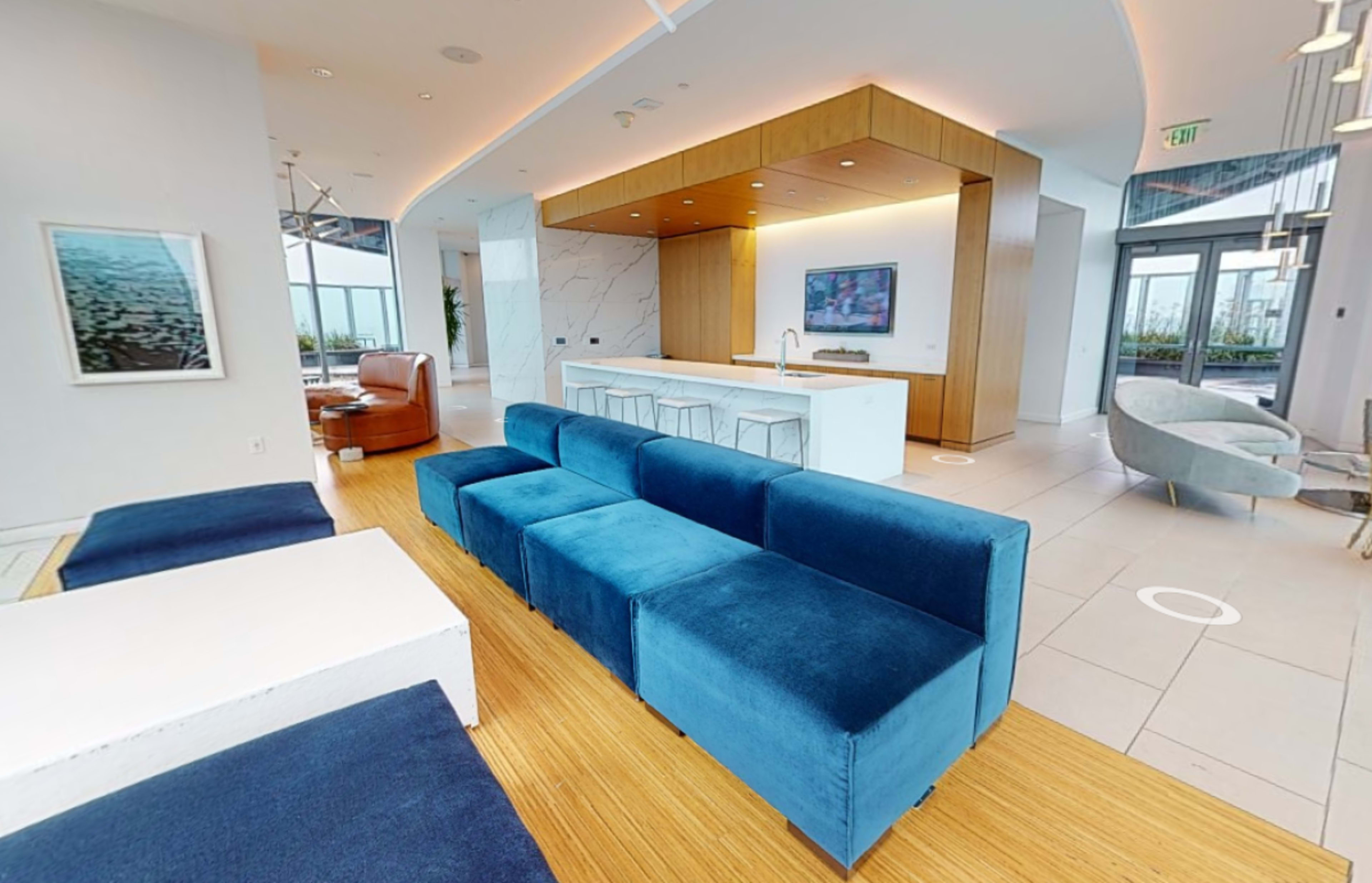 A modern lounge area featuring a blue sectional sofa, a white coffee table, and a kitchen counter in a bright, open space with large windows.