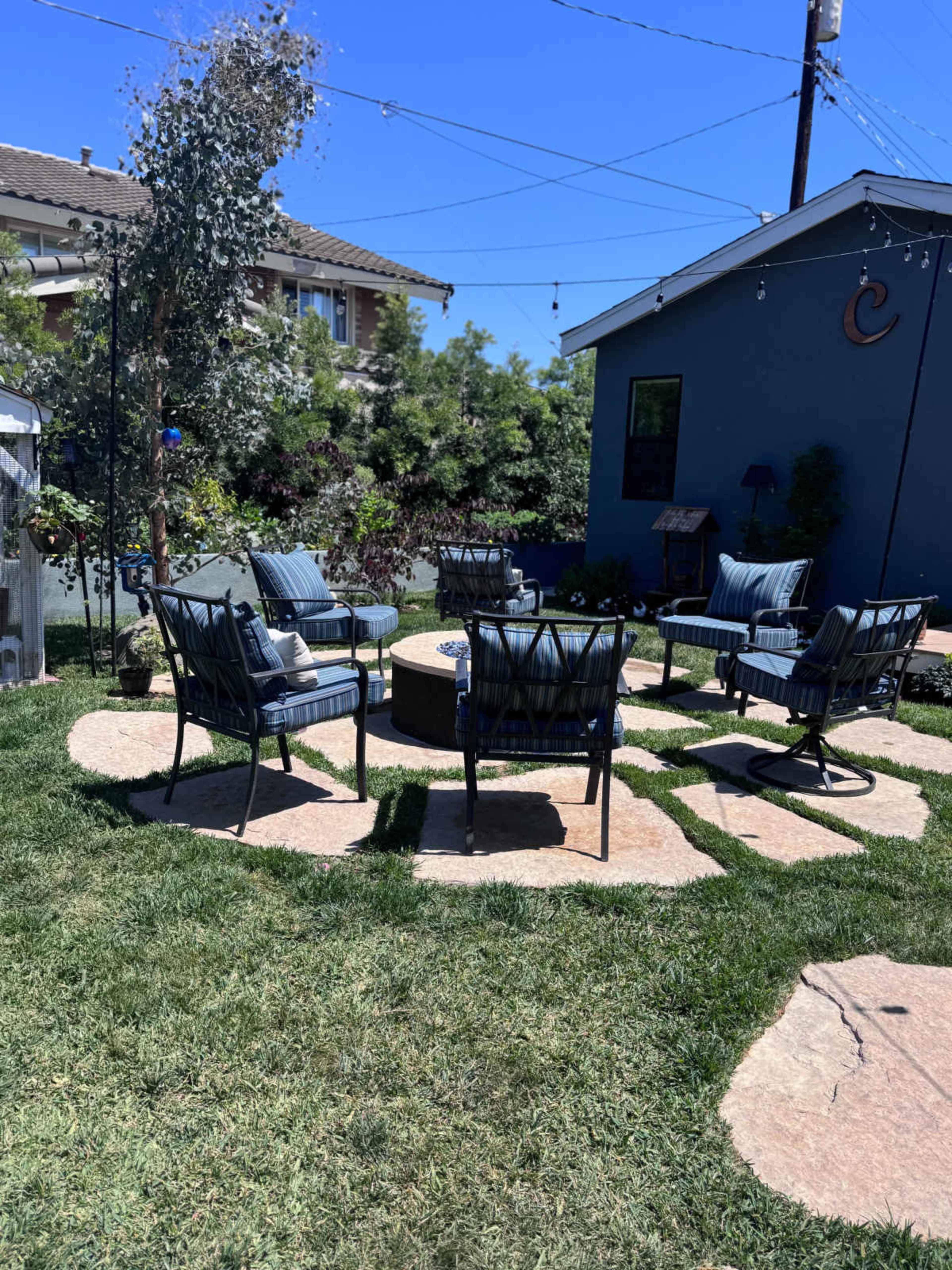 The image shows a backyard patio area with four chairs arranged around a circular stone fire pit, surrounded by green grass and a blue wall in the background.