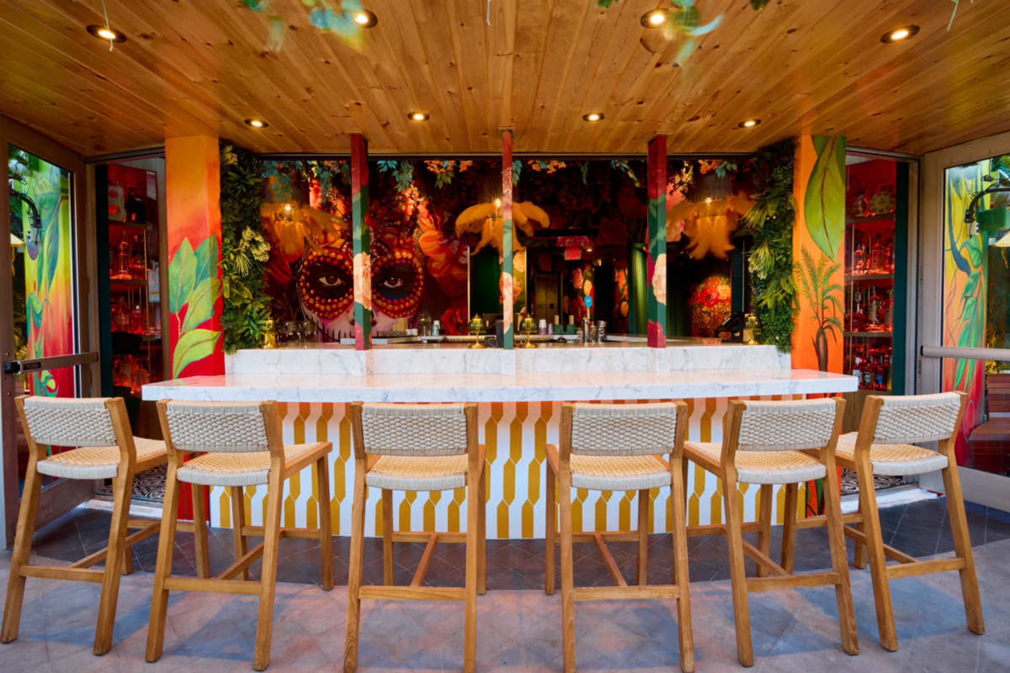 A brightly colored bar with a marble countertop and wooden stools is set against a vibrant mural featuring tropical designs.