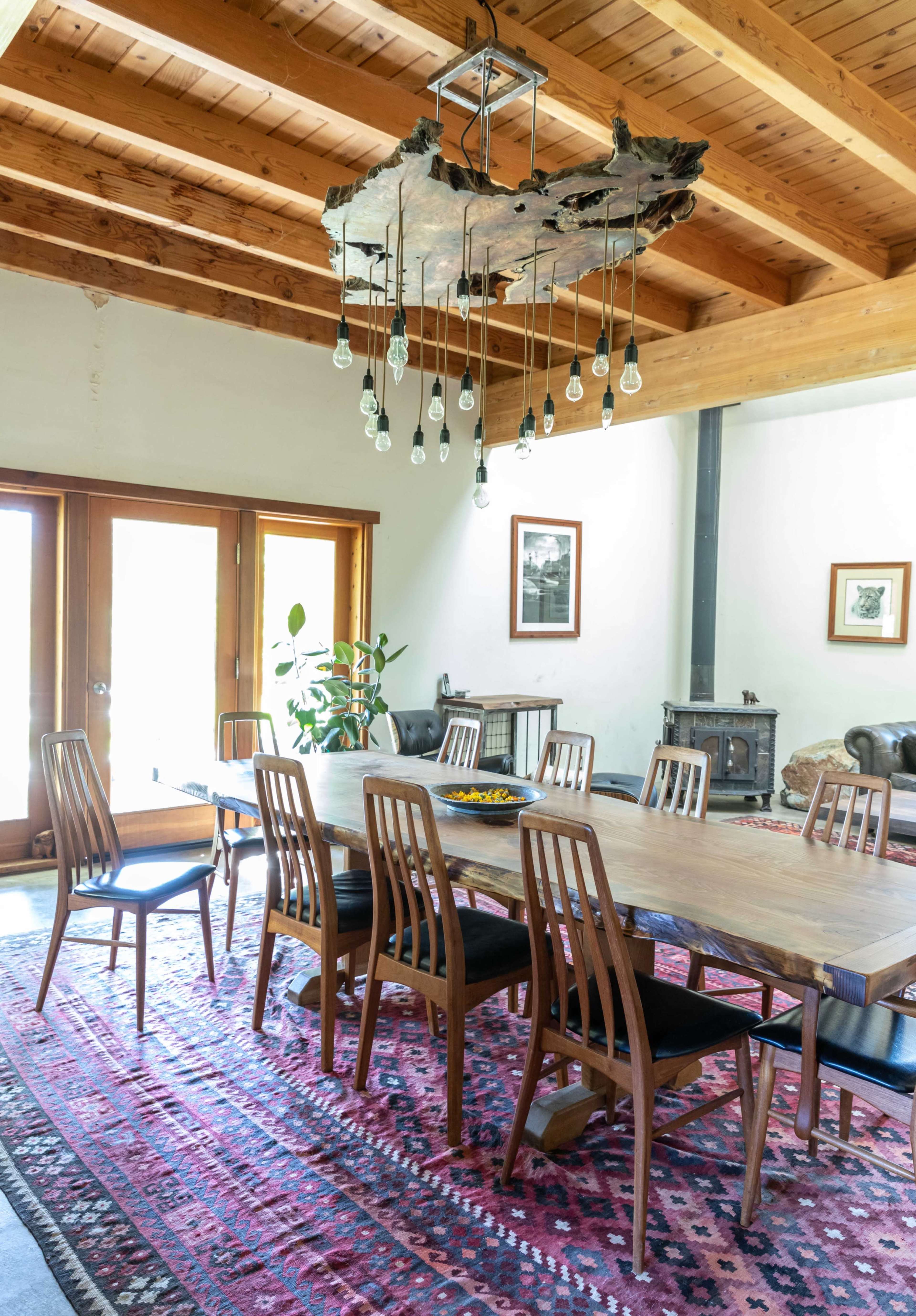 The image shows a spacious dining room featuring a long wooden table surrounded by chairs, with a large chandelier made from wood and light bulbs hanging above, and a rustic wood-burning stove in the corner.