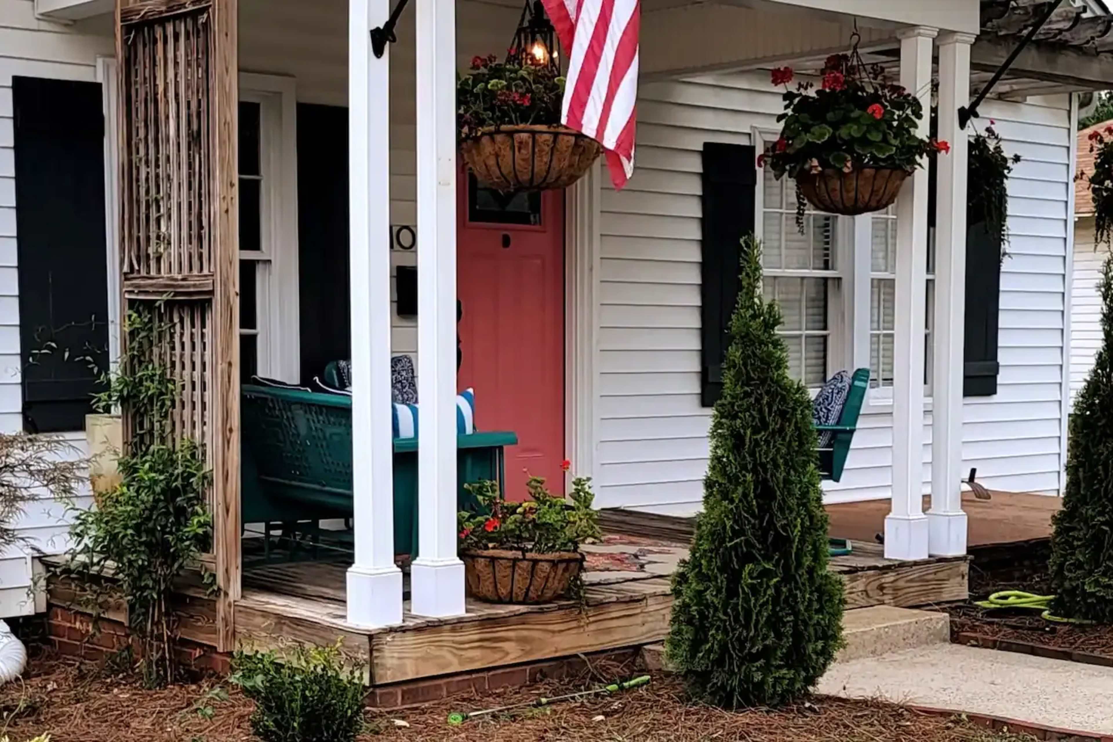 A front porch featuring a pink door, hanging flower baskets, a small evergreen bush, and a couple of chairs.