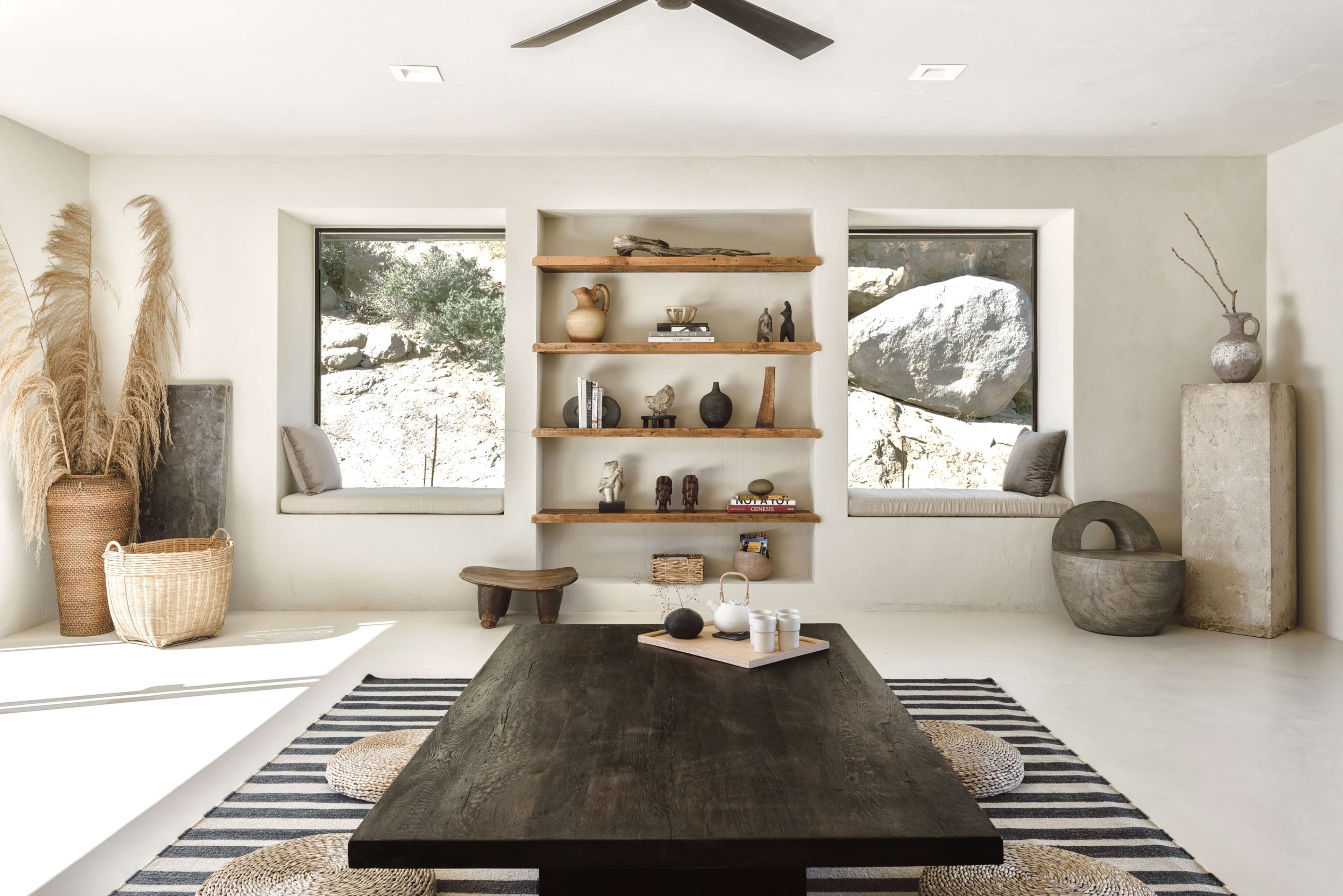 A minimalist room with a large dark wooden table, a striped rug, and shelves displaying various decorative objects, flanked by two windows with window seats.