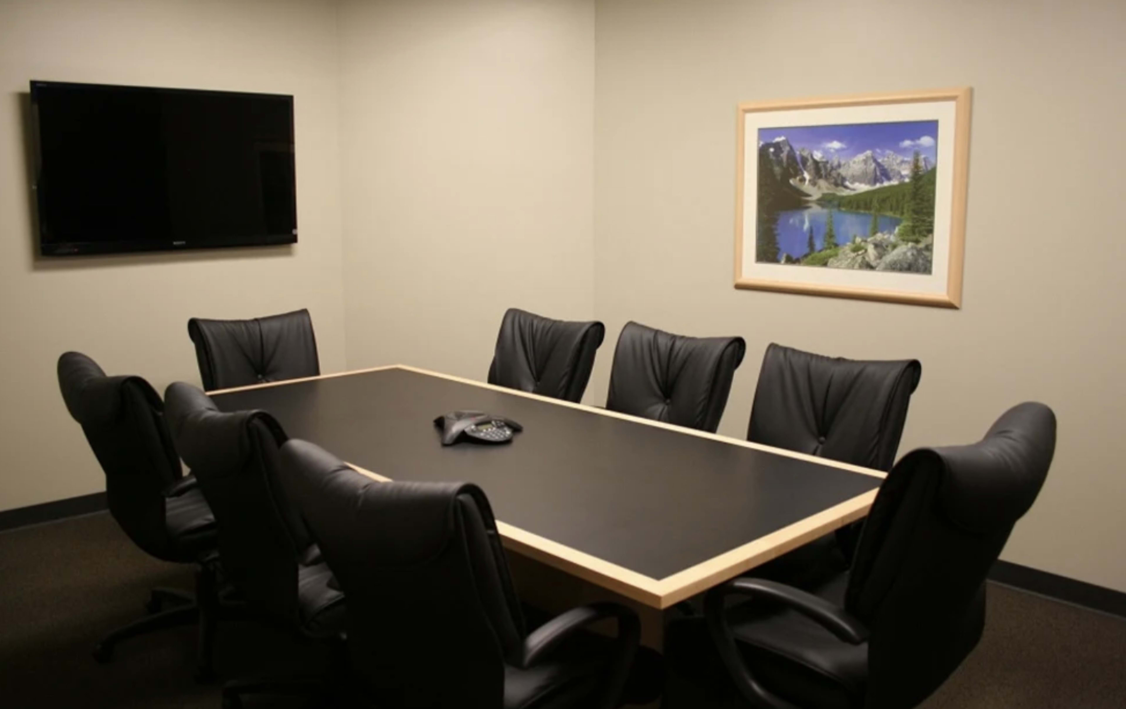 A conference room features a long black table surrounded by eight black chairs, with a mounted television and a framed landscape picture on the wall.