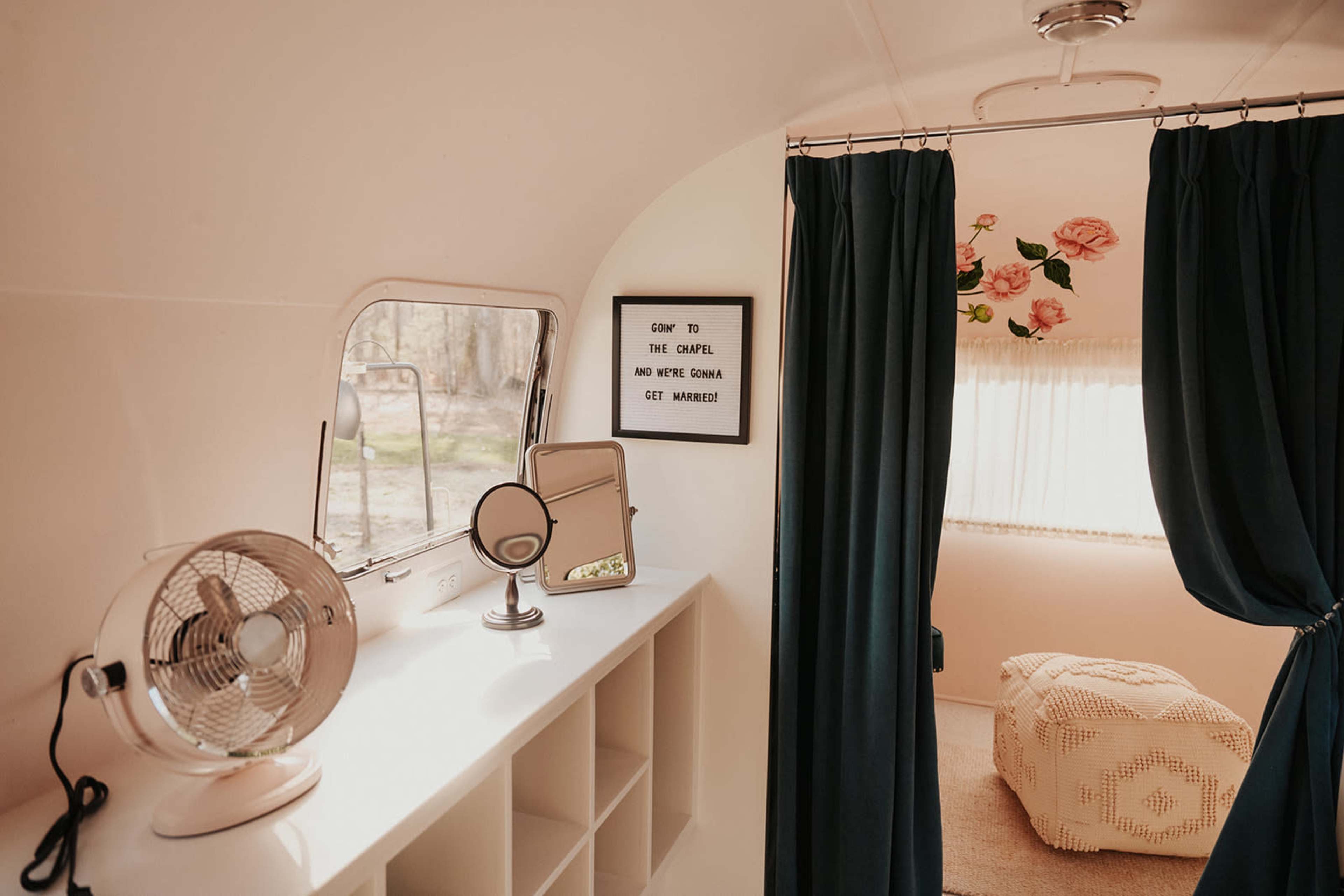 The interior of a vintage trailer featuring a mirror, a fan, and curtains, with a sign on the wall that reads, "Come to the chapel and let's get married."