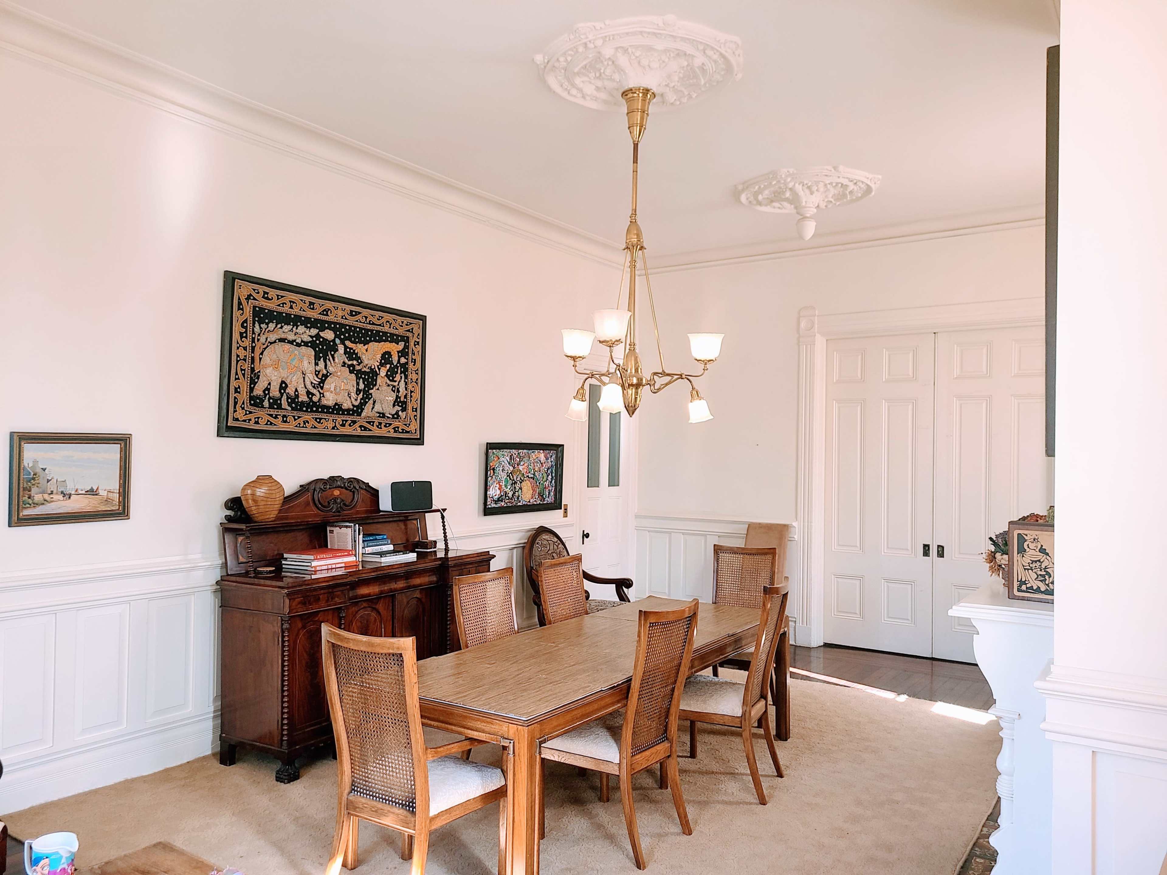 The image shows a formal dining room with a wooden table surrounded by wicker chairs, a chandelier hanging from the ceiling, and framed artwork on the walls.