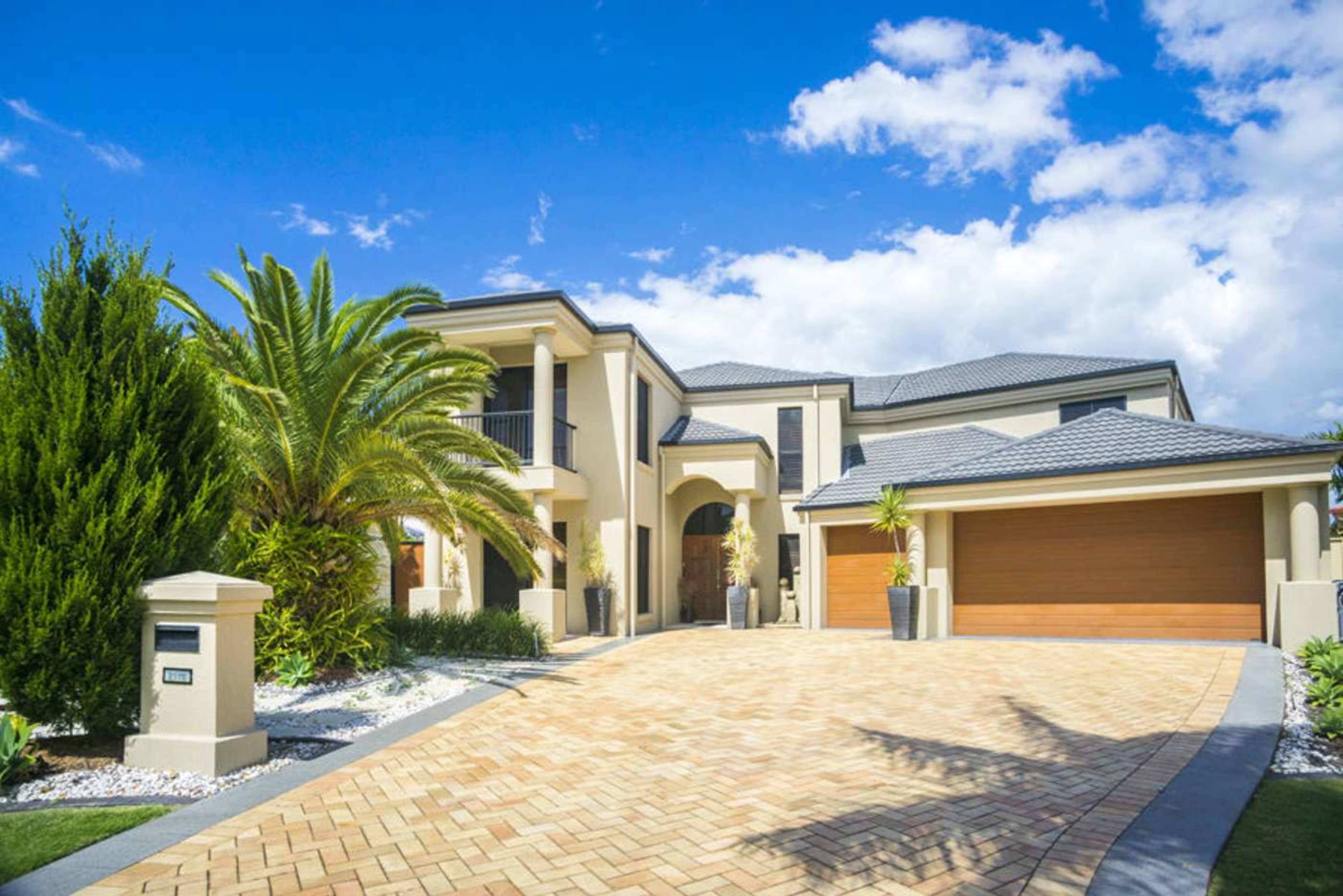 A large, modern two-story house with a paved driveway and palm trees in the front yard.
