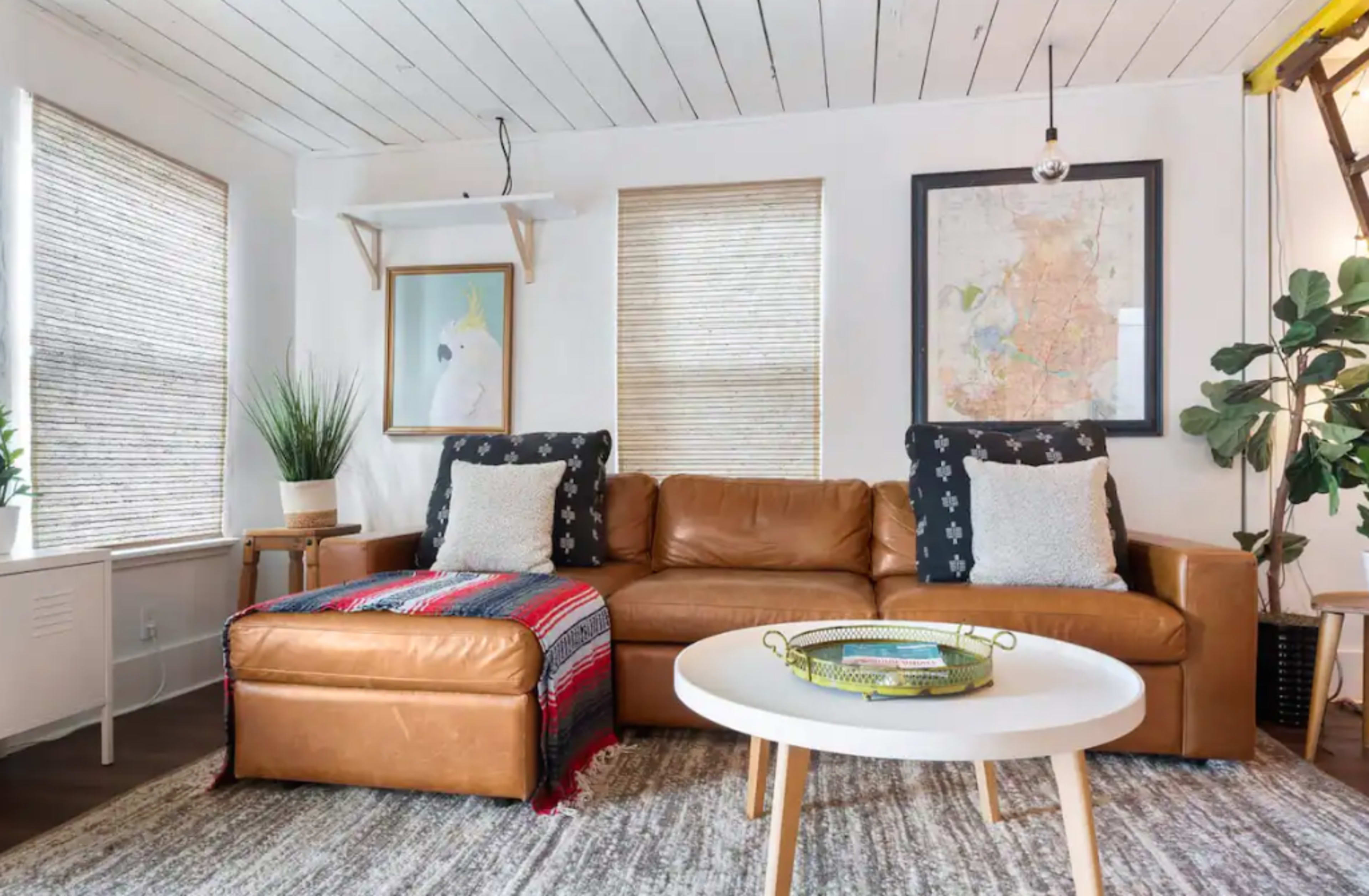 A cozy living room features a brown leather sofa with decorative pillows, a round coffee table, and natural light streaming through window shades.