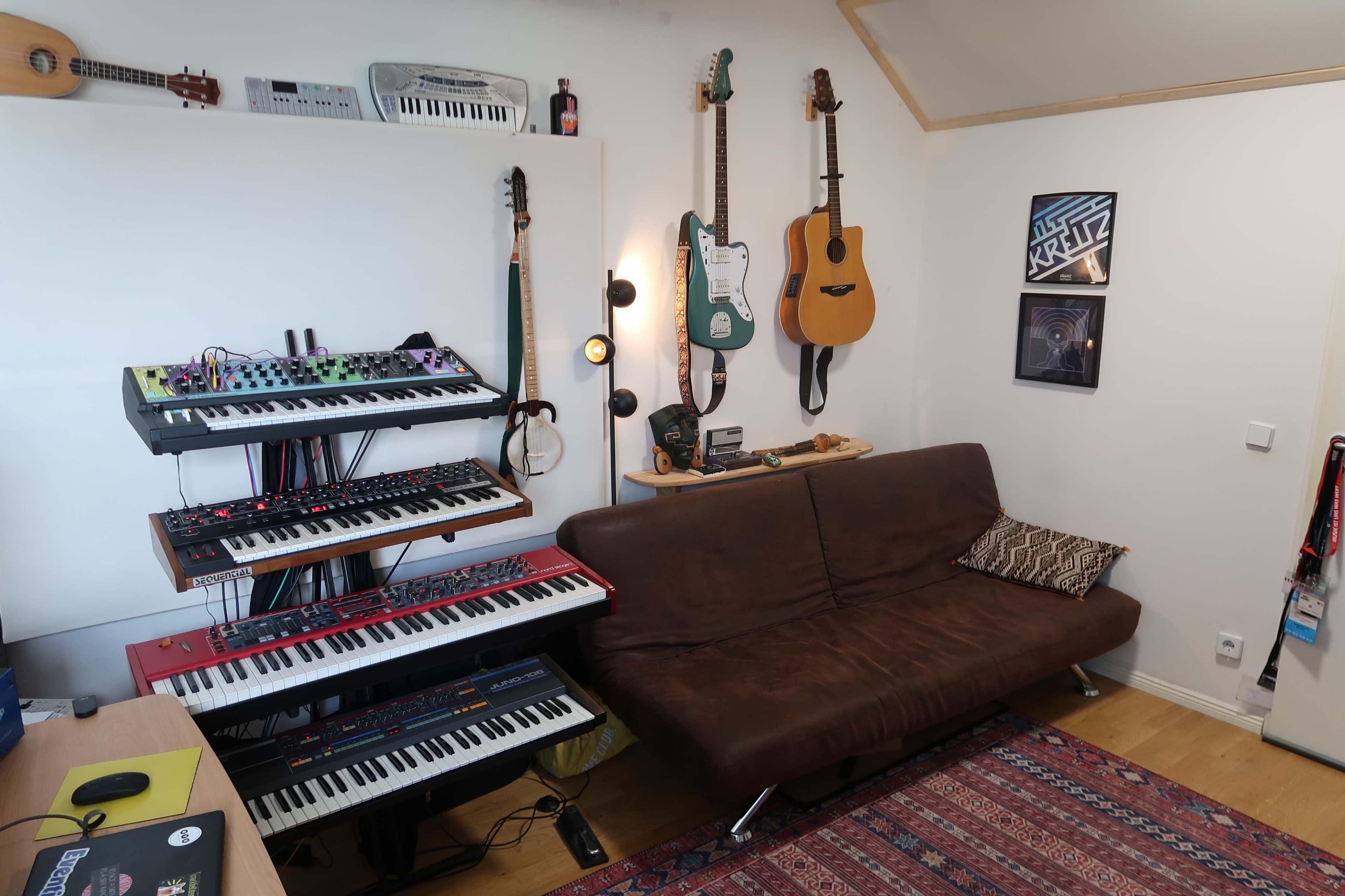 The image shows a music studio setup featuring a couch, synthesizers, and several guitars mounted on the wall.