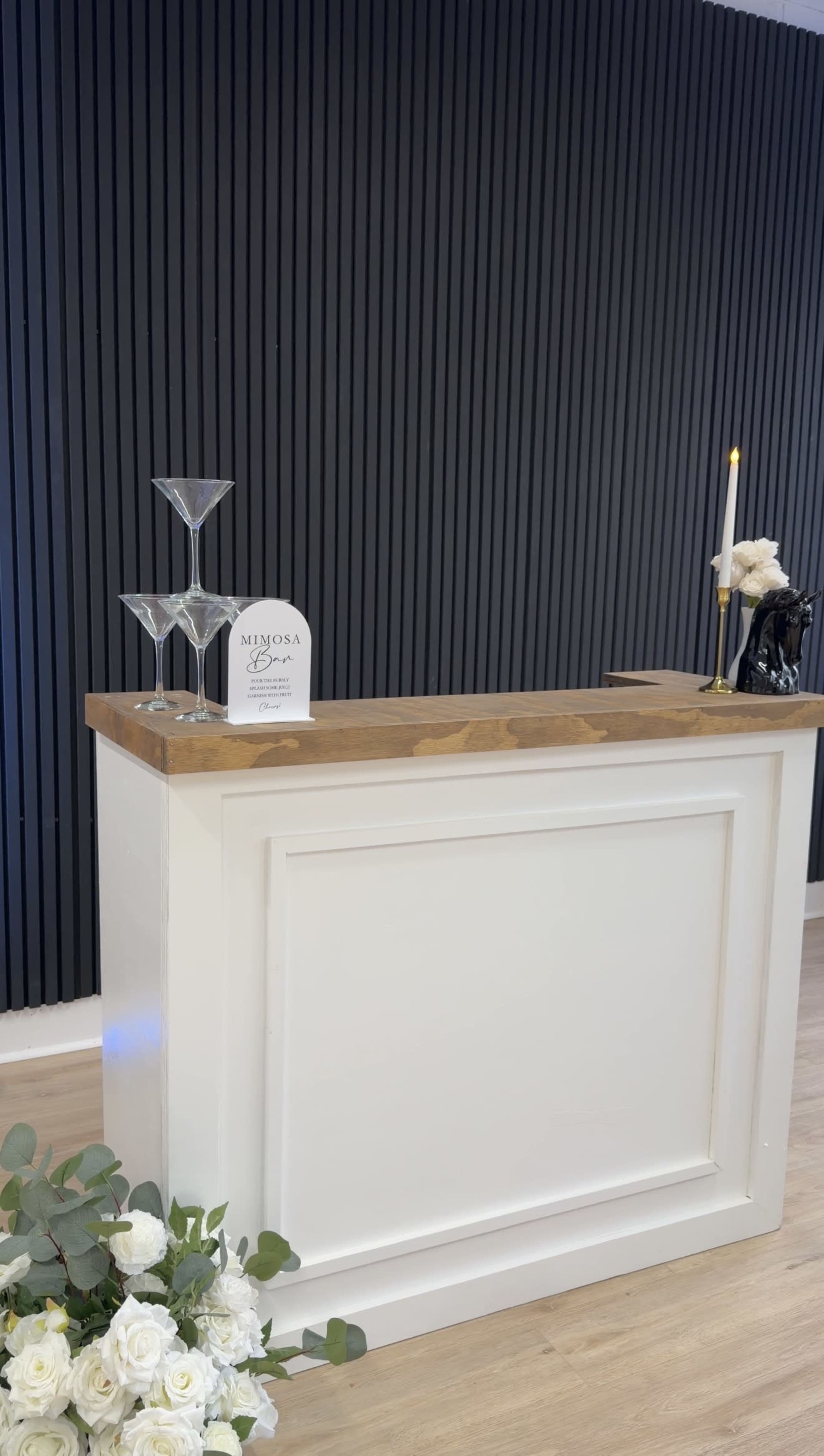 A white bar counter with a wooden top is set against a black wall, adorned with a few glassware and a candle, accompanied by white flowers in the foreground.