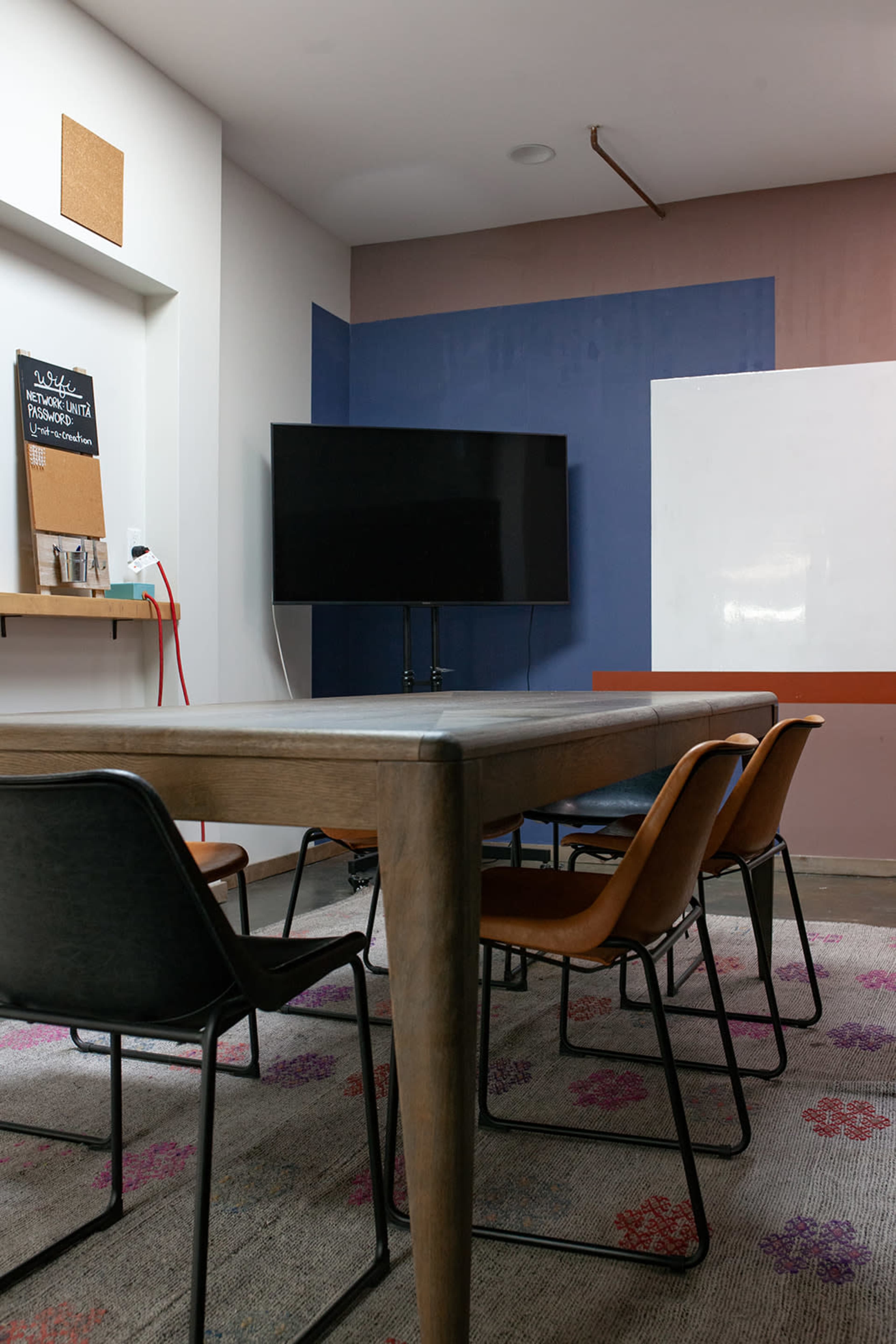 The image shows a modern conference room featuring a long wooden table surrounded by black and brown chairs, a large television mounted on the wall, and a whiteboard.