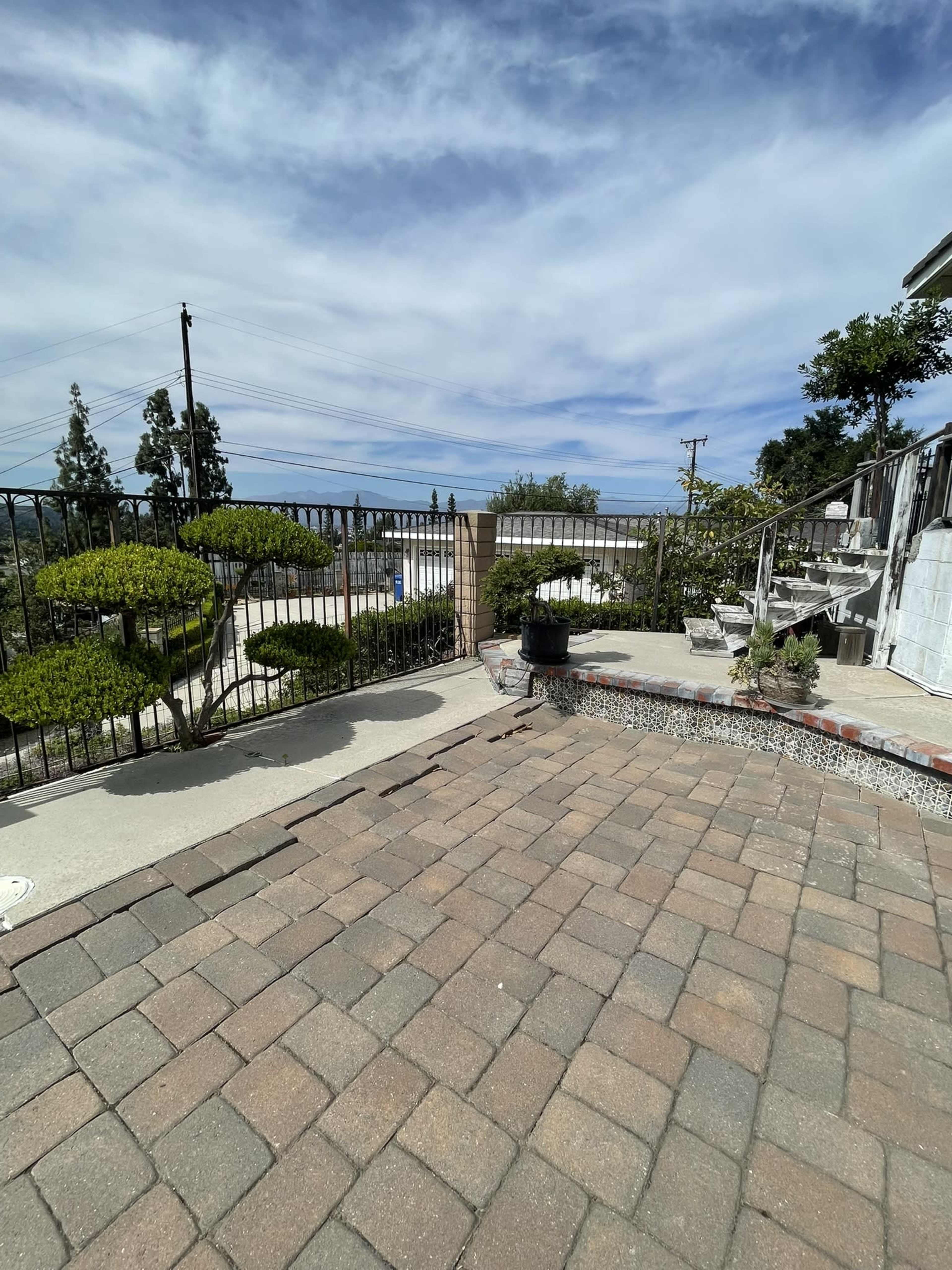 The image shows a landscaped patio area with patterned brick flooring, bordered by a wrought iron fence and potted plants, overlooking a distant view under a partly cloudy sky.