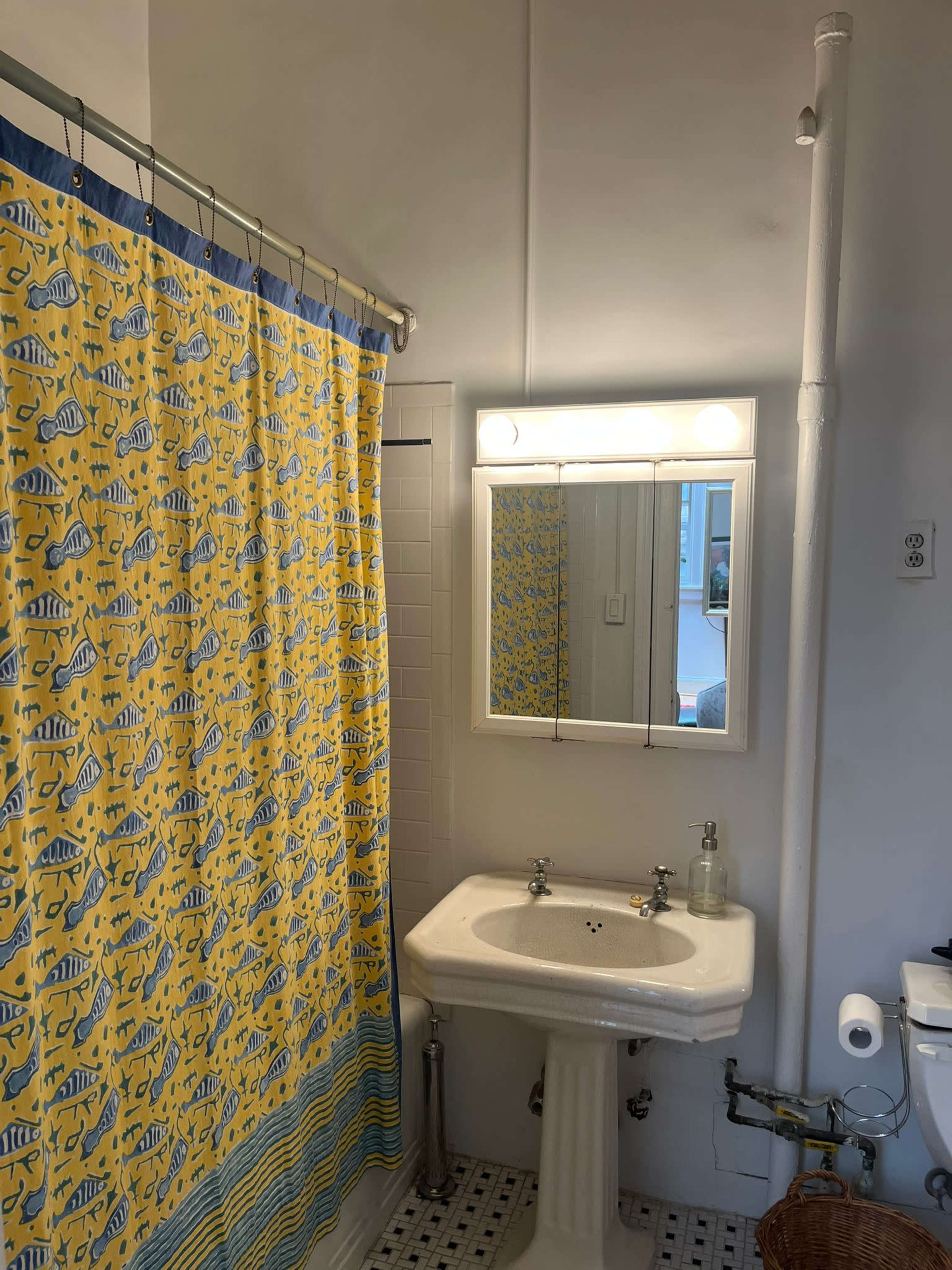 The image shows a bathroom featuring a white pedestal sink, a mirror above it, and a shower curtain with a yellow fish pattern.