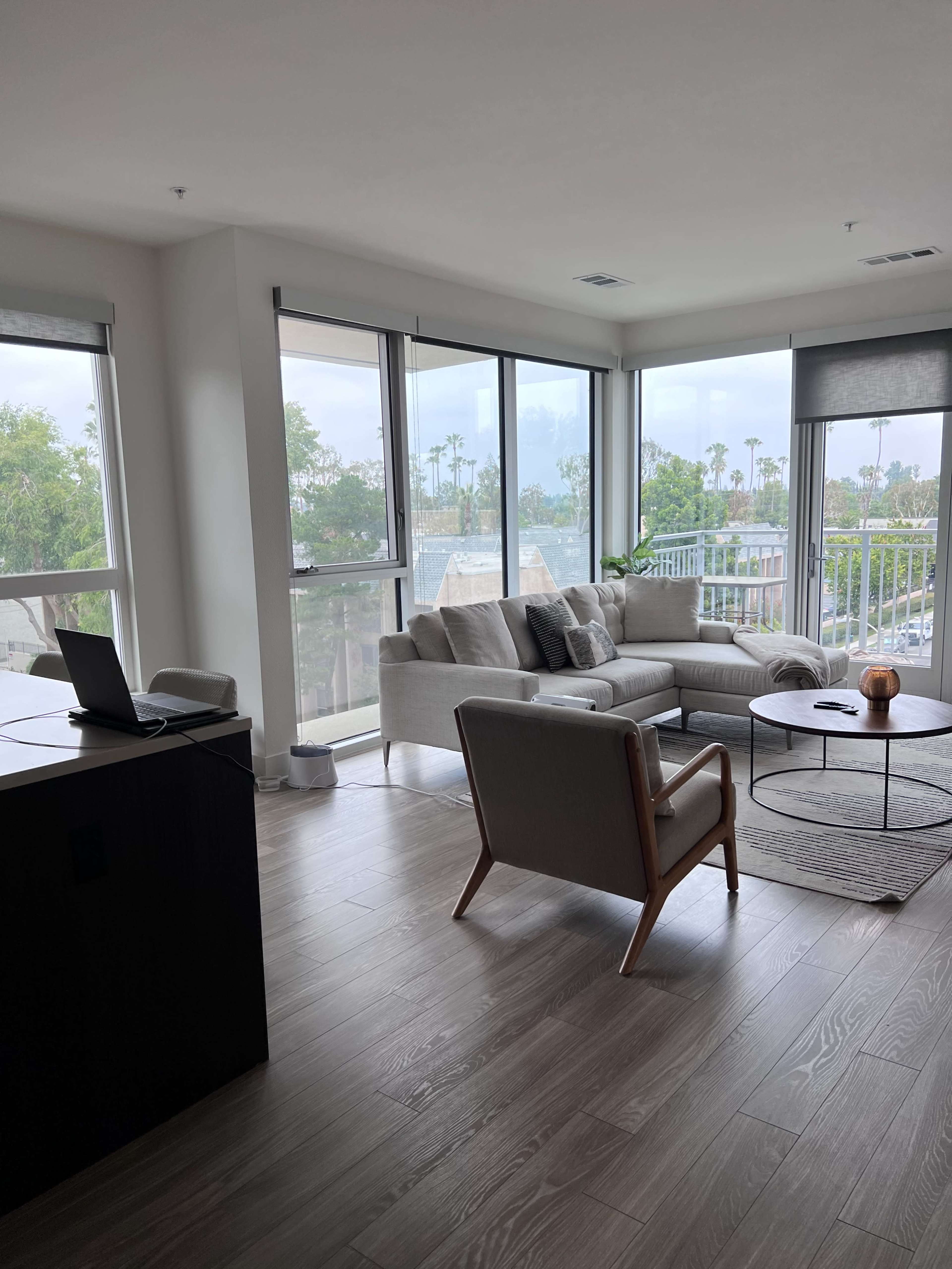 A modern living room features a light-colored sofa, a round coffee table, and large windows overlooking greenery and palm trees.