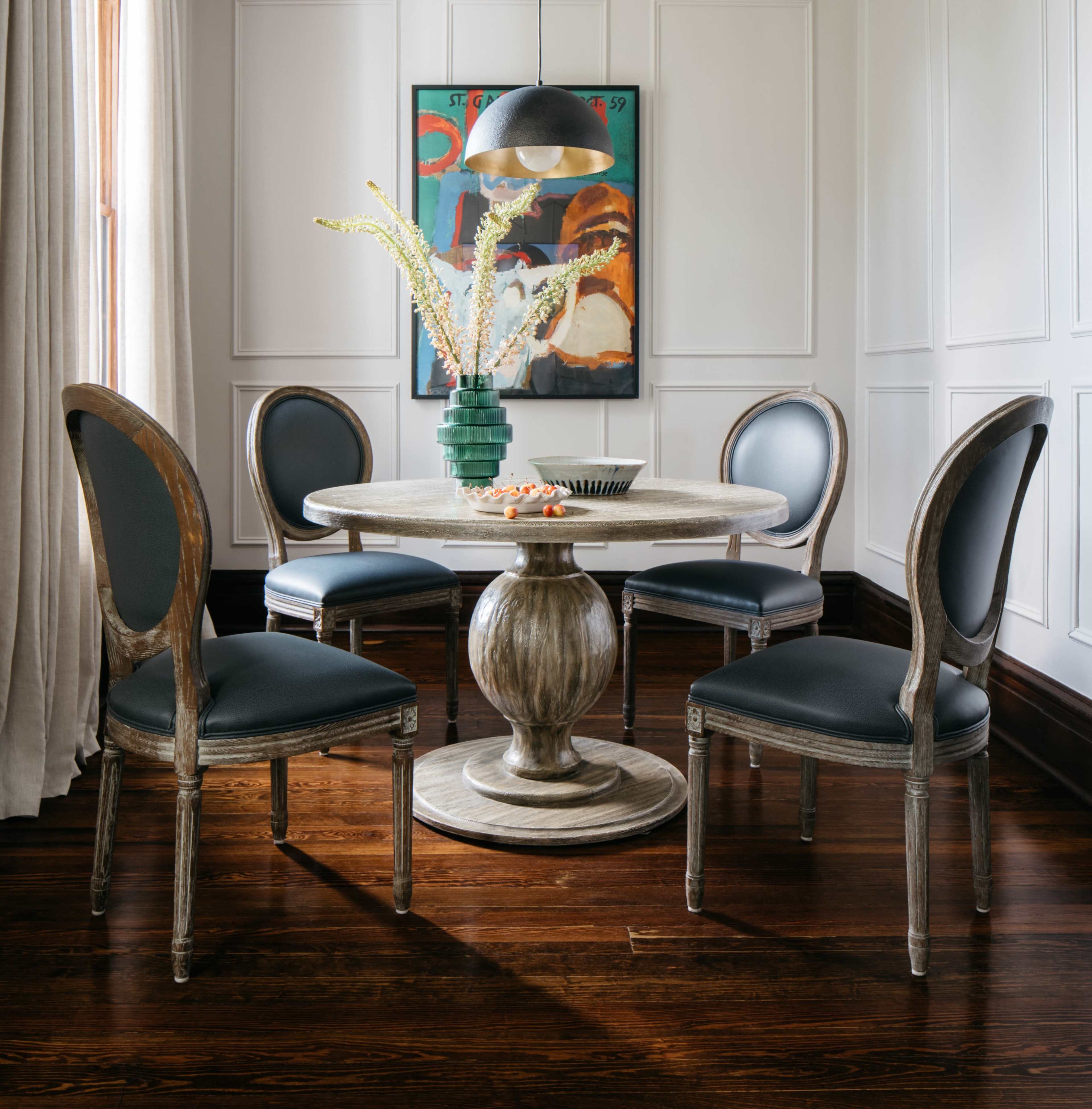 A round dining table is set in a well-lit room with four upholstered chairs, a pendant light above, and a framed artwork on the wall.