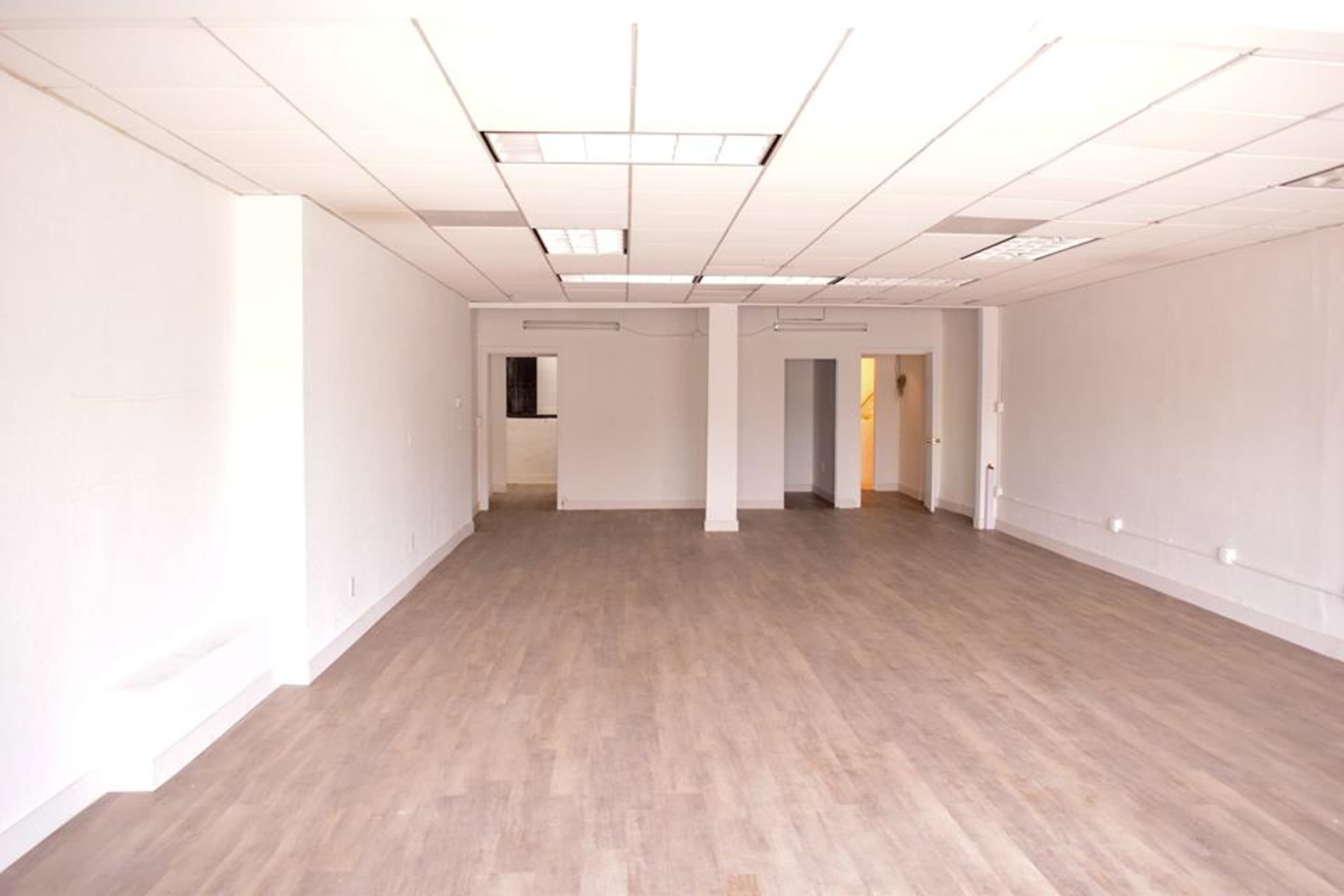 A spacious, empty room with wood-like flooring and white walls, featuring multiple doorways and fluorescent lighting.
