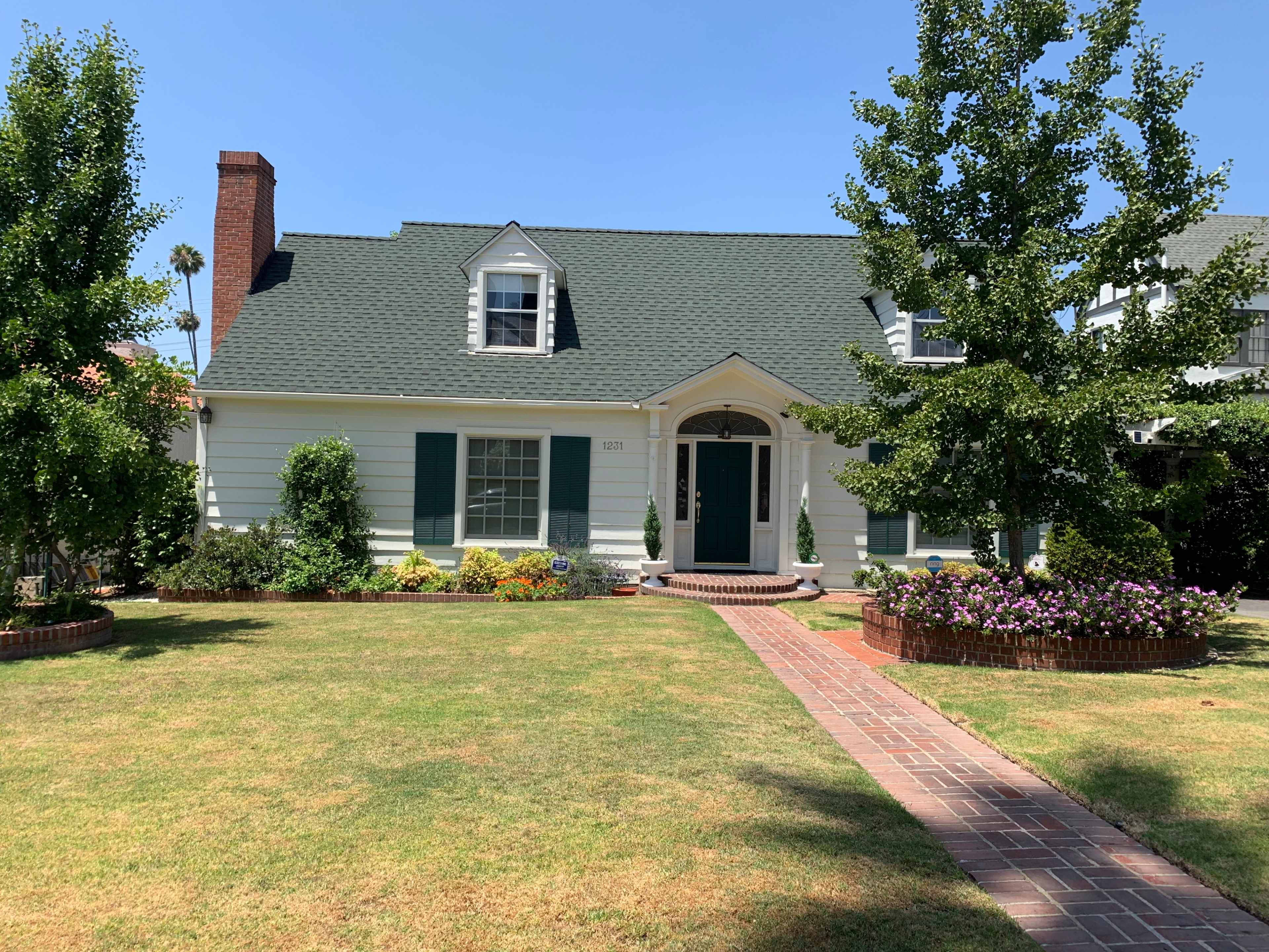 A light-colored two-story house with a green roof features a front pathway lined with flowers and shrubs.