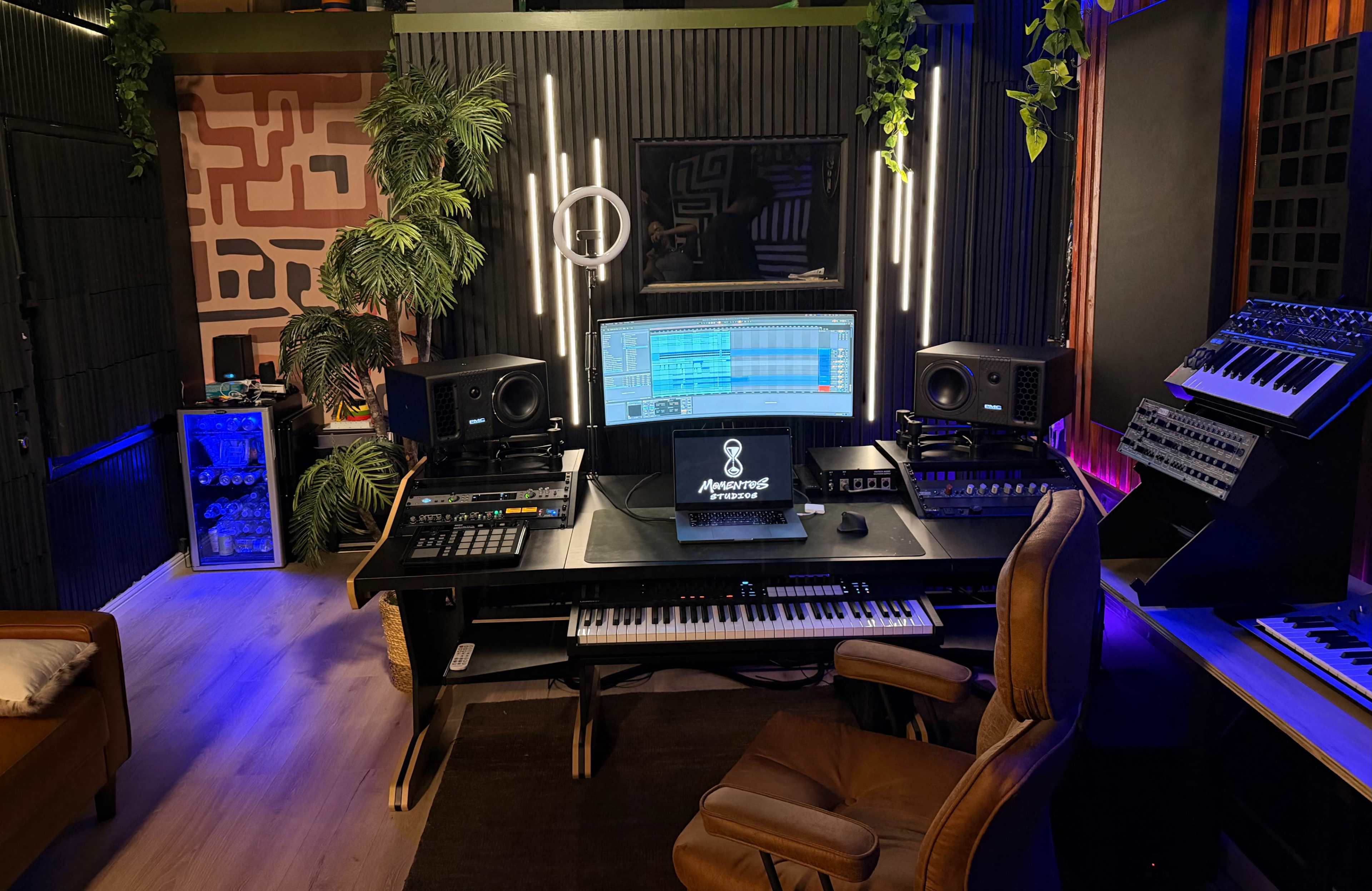 The image shows a modern music studio featuring a central workstation with a computer, a keyboard, speakers, and various music equipment, surrounded by decorative plants and ambient lighting.