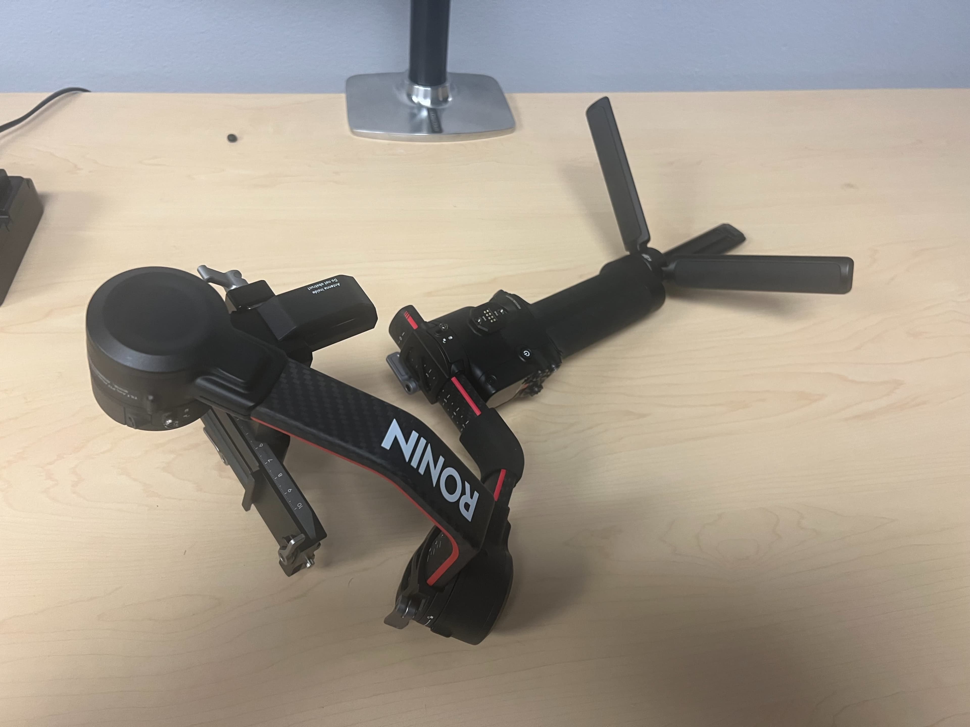 The image shows a gimbal stabilizer with a camera mount, placed on a wooden table, alongside its propeller attachment.