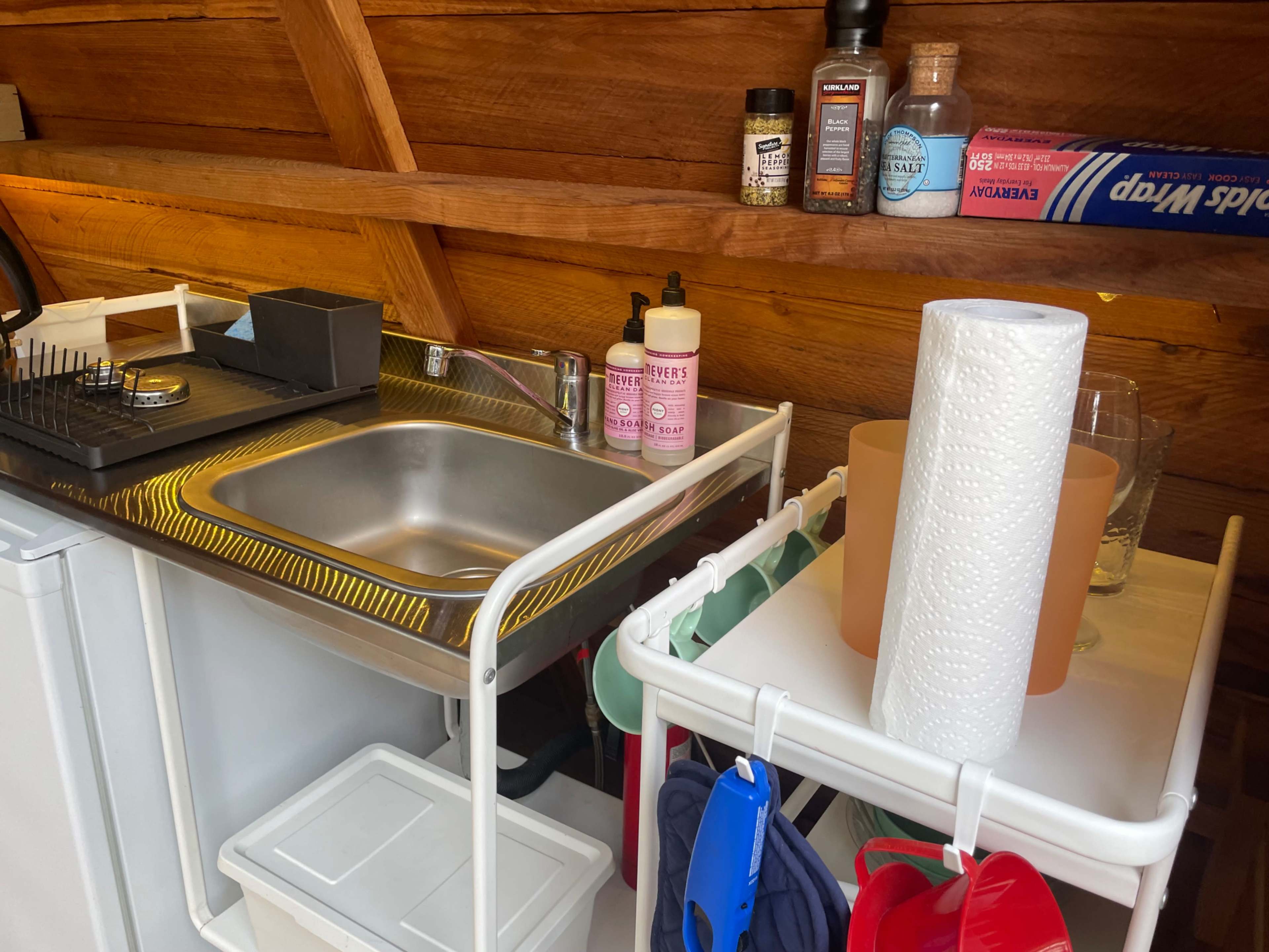 A small kitchen area with a stainless steel sink, a portable gas stove, and various items organized on shelves, including condiments and paper towels.