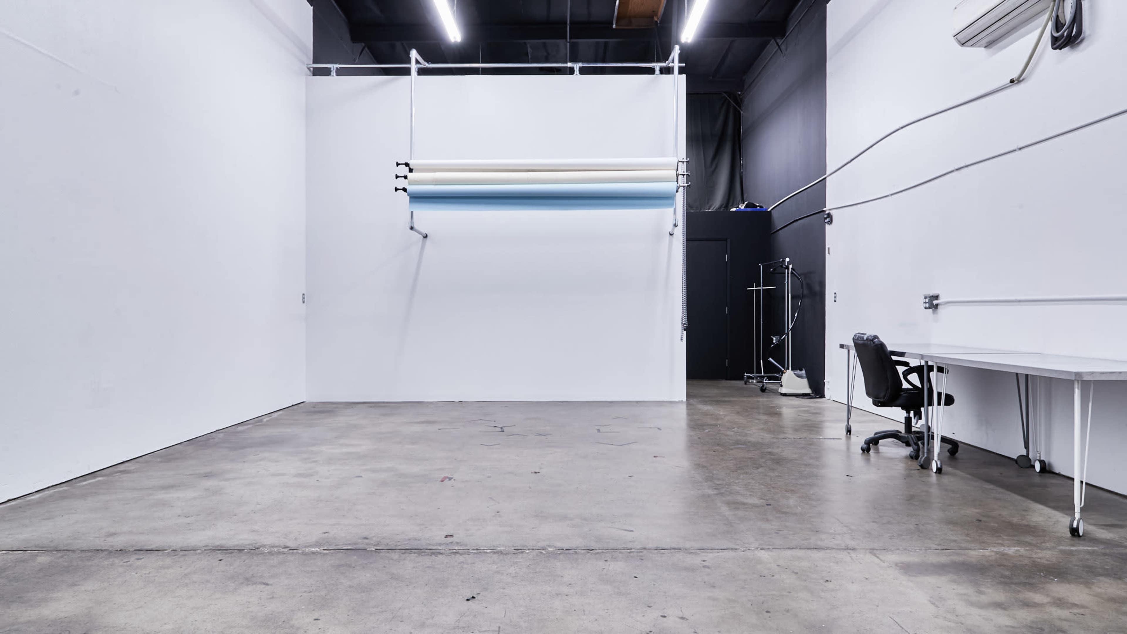 A spacious studio with a concrete floor, bare white walls, and a large backdrop holder mounted on the wall, alongside a desk and a chair.