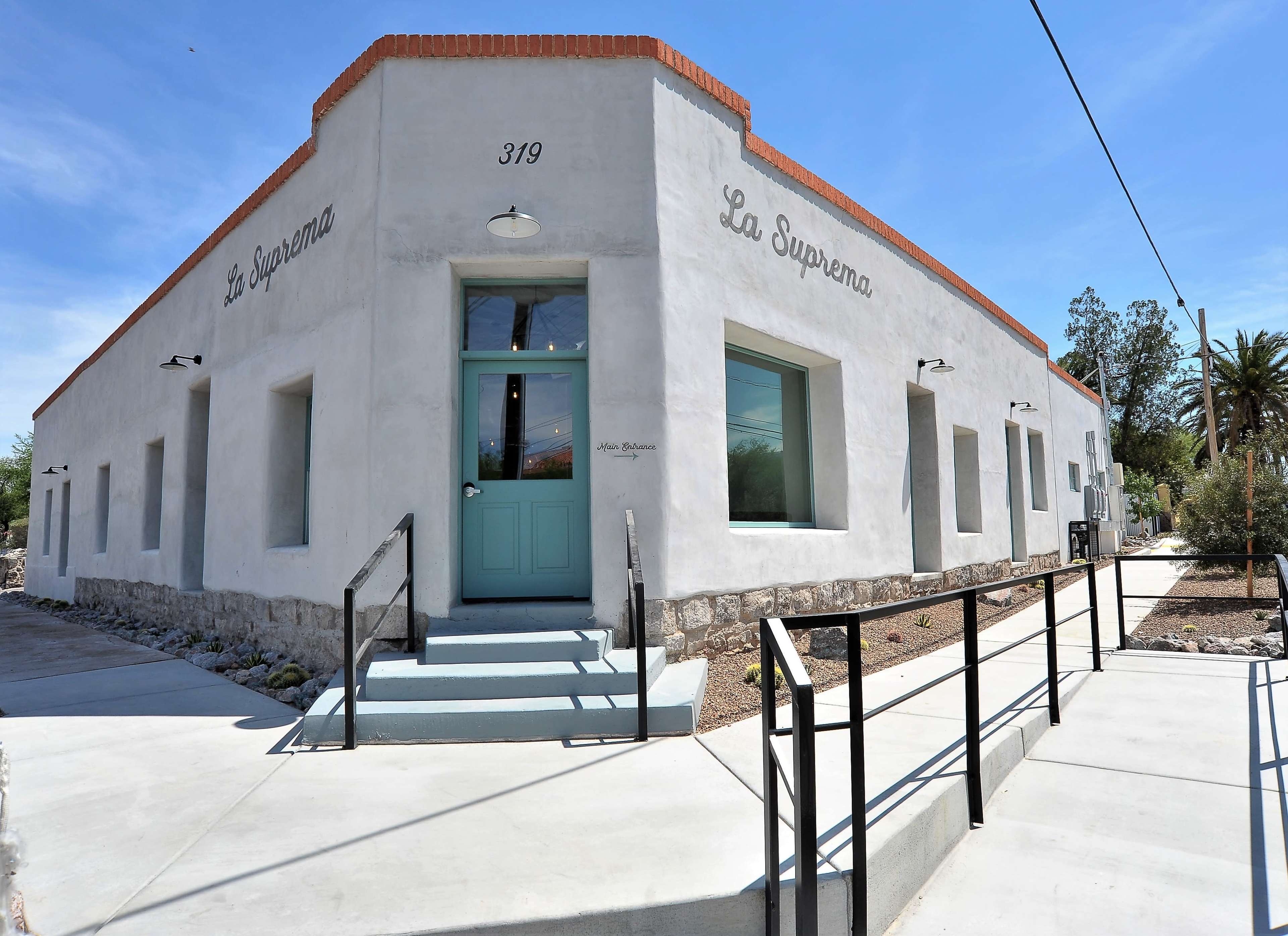 A modern, single-story building with a light-colored exterior labeled "La Suprema" at 319, featuring large windows and a set of concrete steps leading to a teal door.