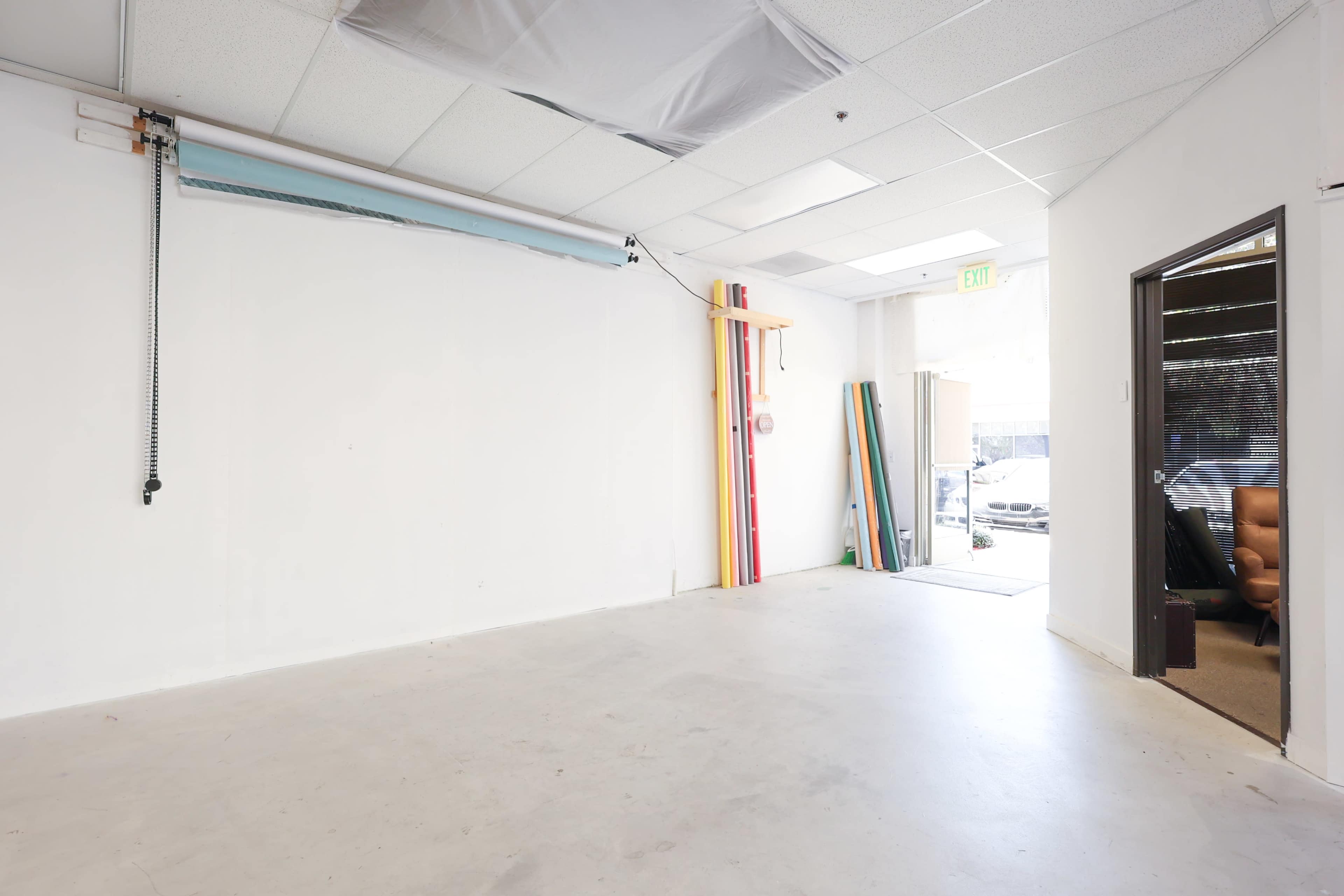 The image shows an empty room with white walls, concrete flooring, and a doorway leading to an outdoor area, along with colorful panels stacked against the wall.