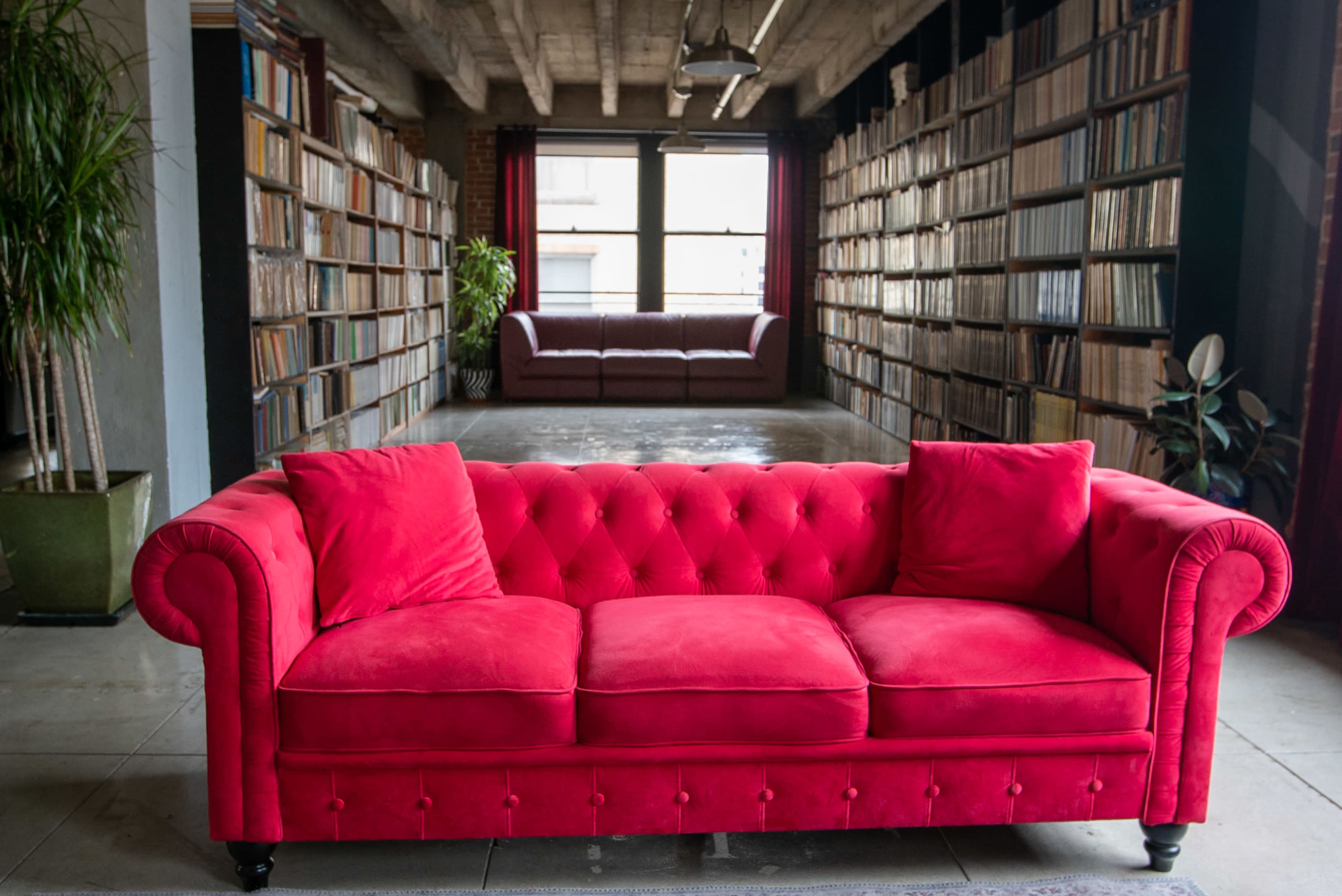 A red tufted couch is positioned in the foreground, facing a room with two rows of bookshelves and another couch in the background.