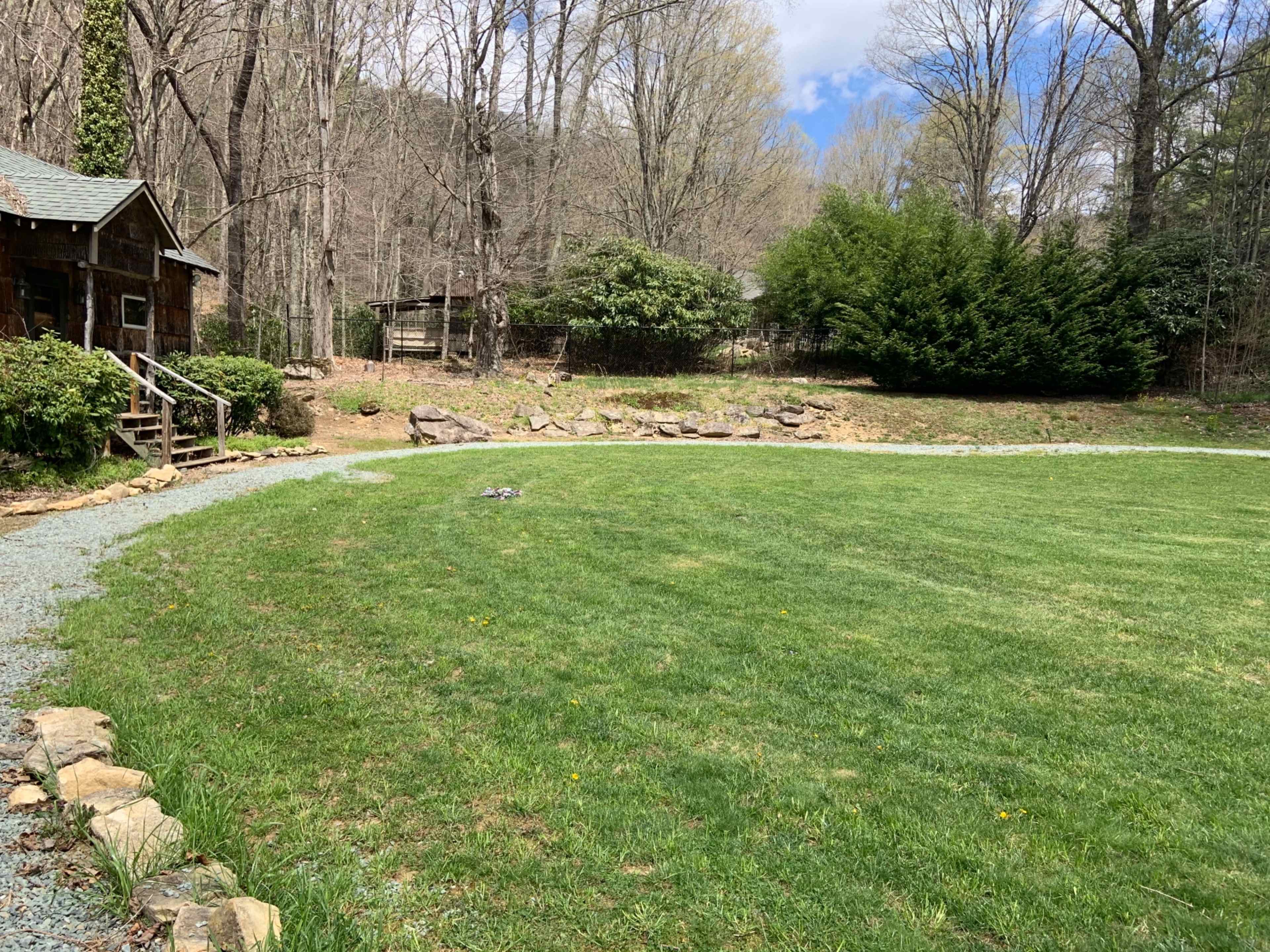 A grassy area with a stone border curves around a wooden cabin, surrounded by trees and shrubs.