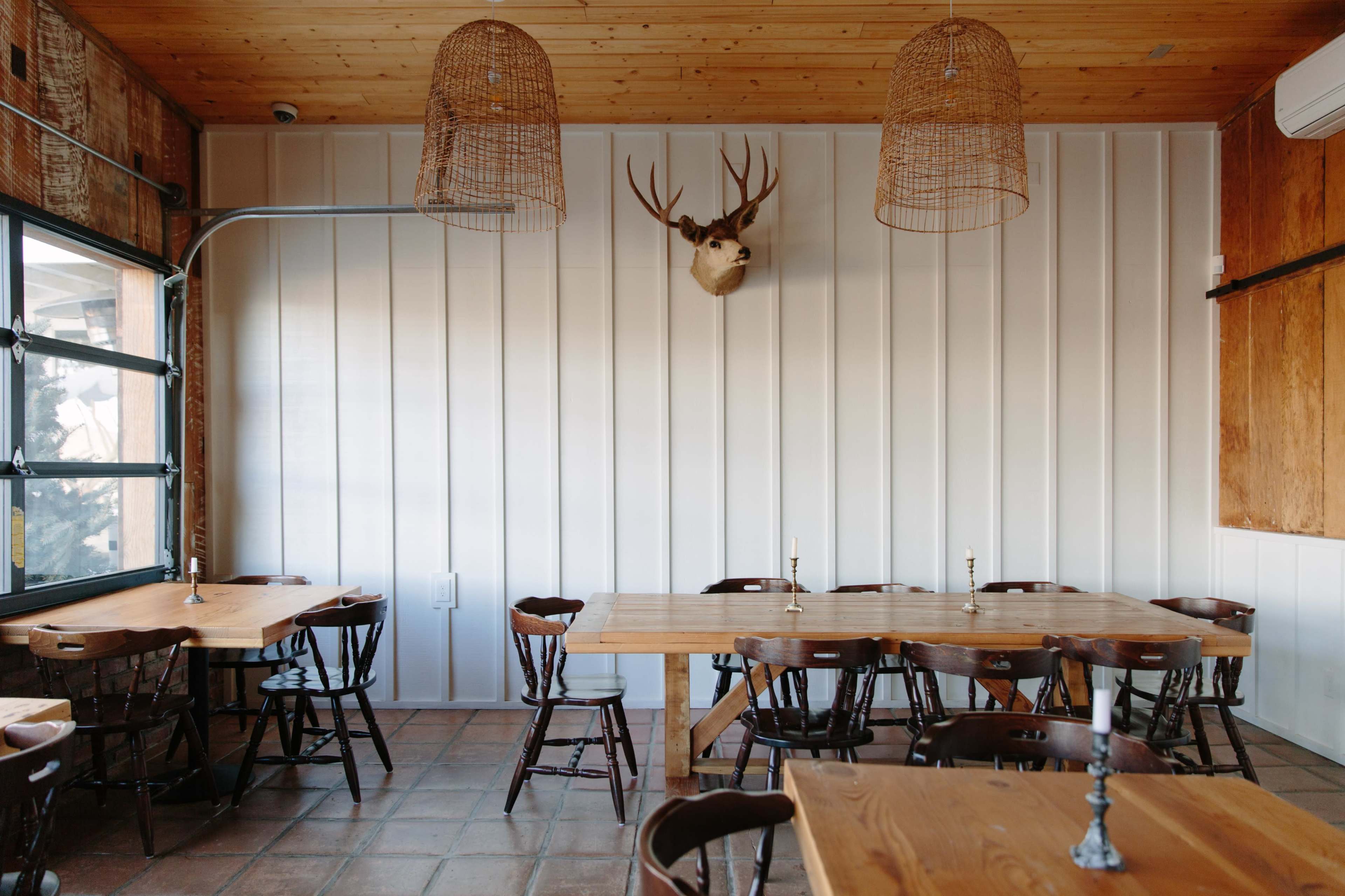A spacious dining area features wooden tables, chairs, and a mounted deer head on the wall.