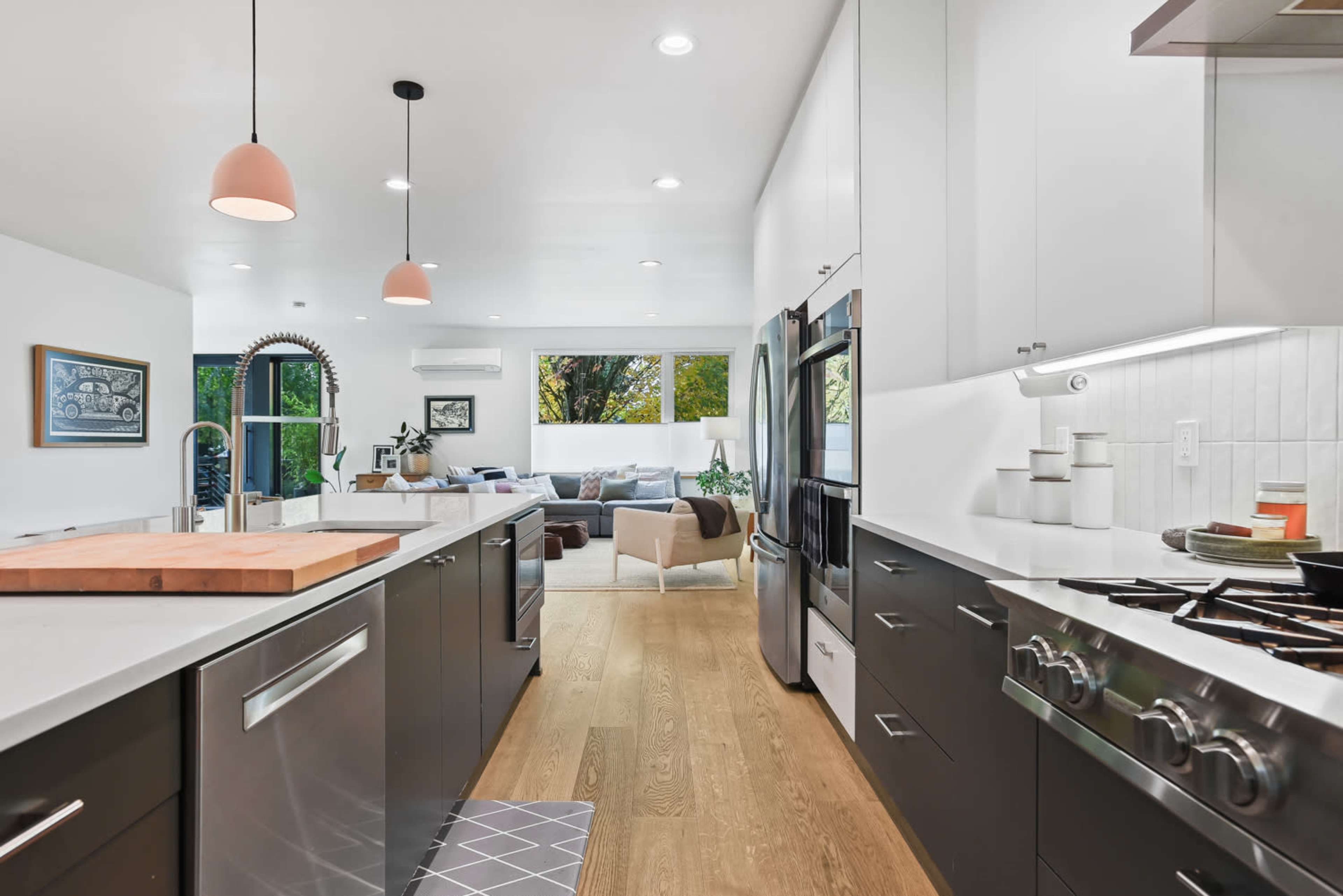 A modern kitchen features dark cabinets, light countertops, and stainless steel appliances, leading into a bright living area with a sofa and large windows.