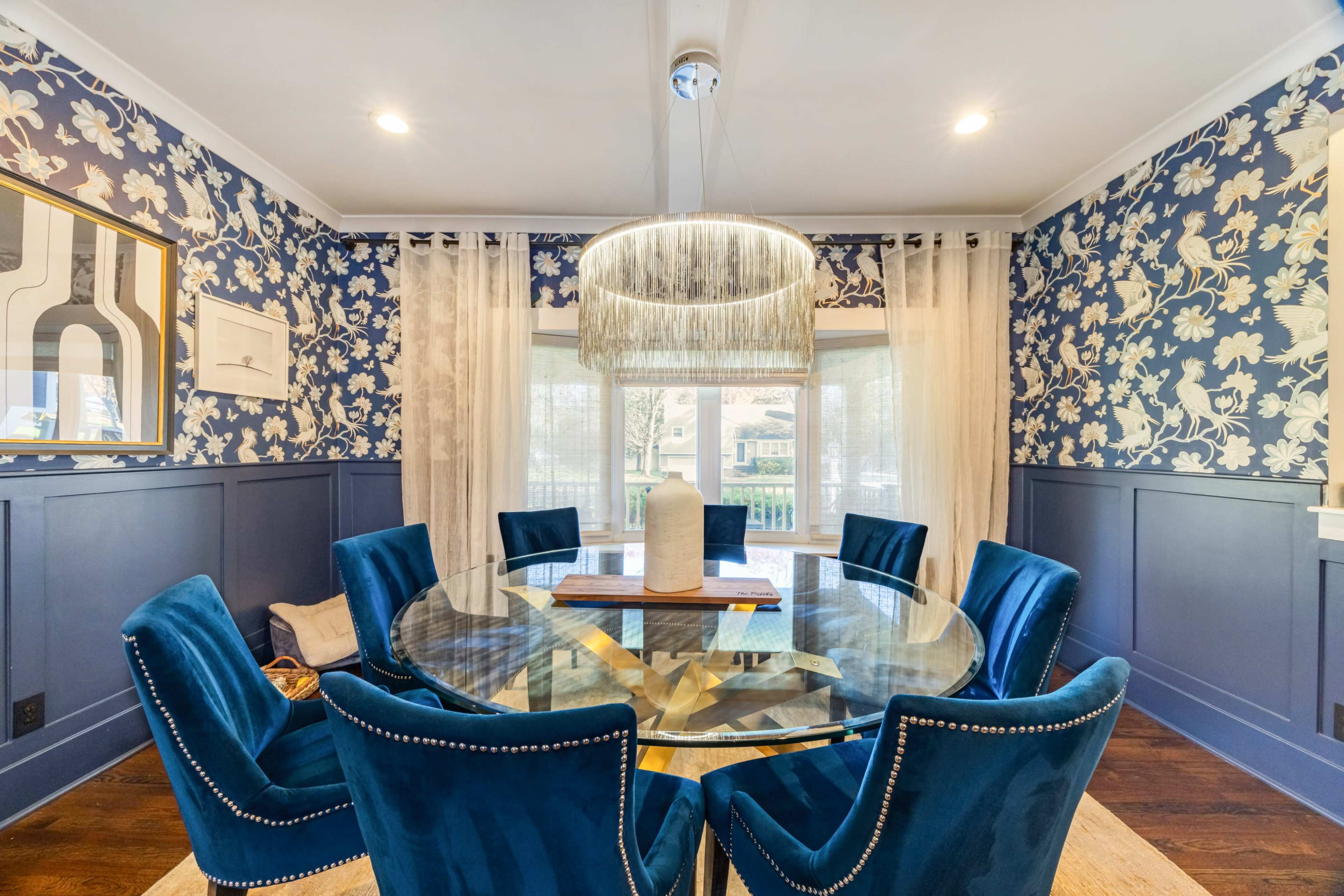 A round glass dining table surrounded by blue upholstered chairs sits in a room with floral wallpaper and large windows.